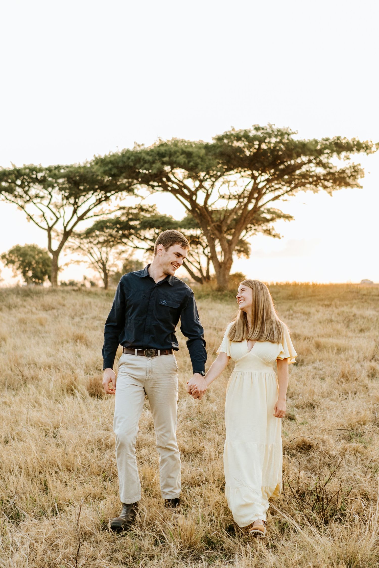 Happy couple holding hands walking through dry golden savanna grassland with acacia trees at sunset.