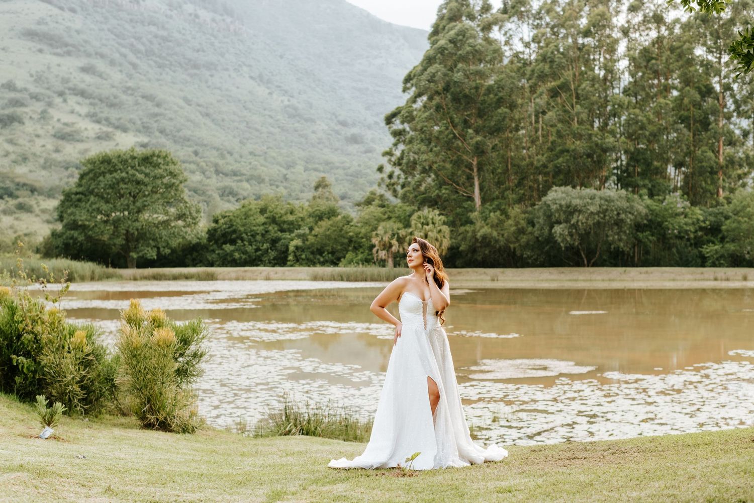 Bride in white gown with slit poses gracefully near serene lake with lush mountain backdrop.