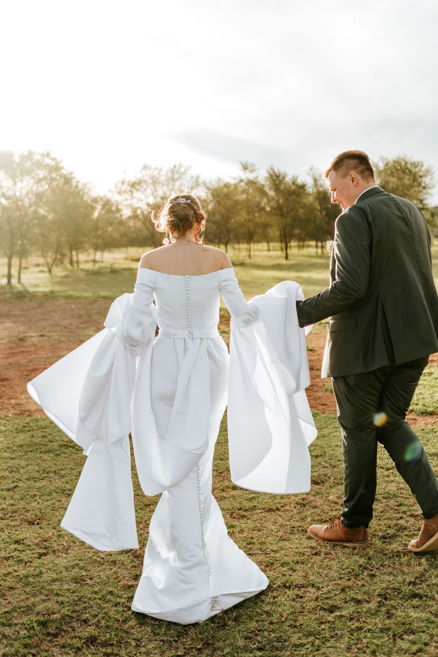 Bride in off-shoulder white gown with dramatic sleeves walks with groom through scenic outdoor field.