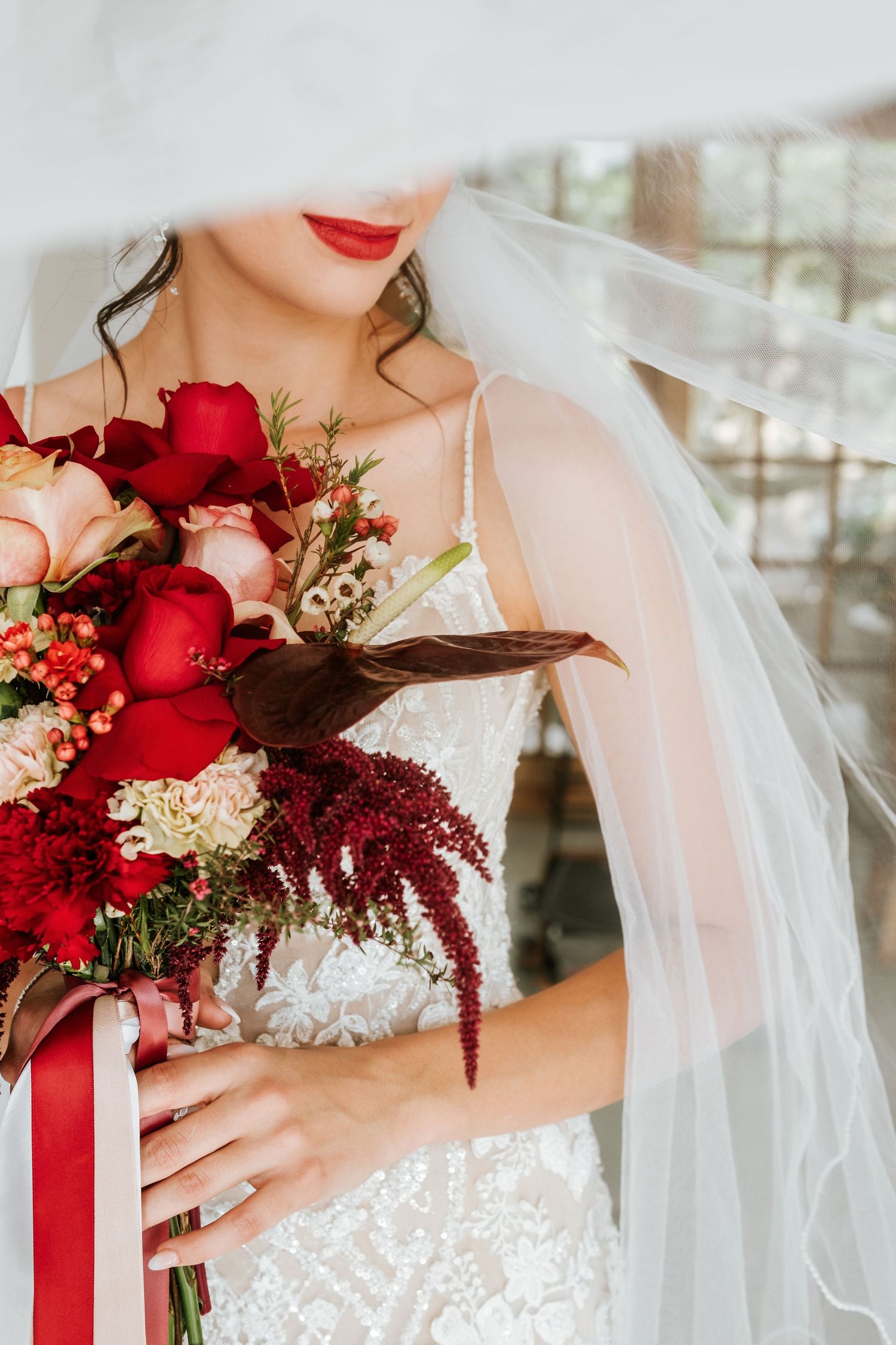 Bride in lace gown holding a stunning red and pink bouquet with roses and amaranthus, wearing a flowing veil.