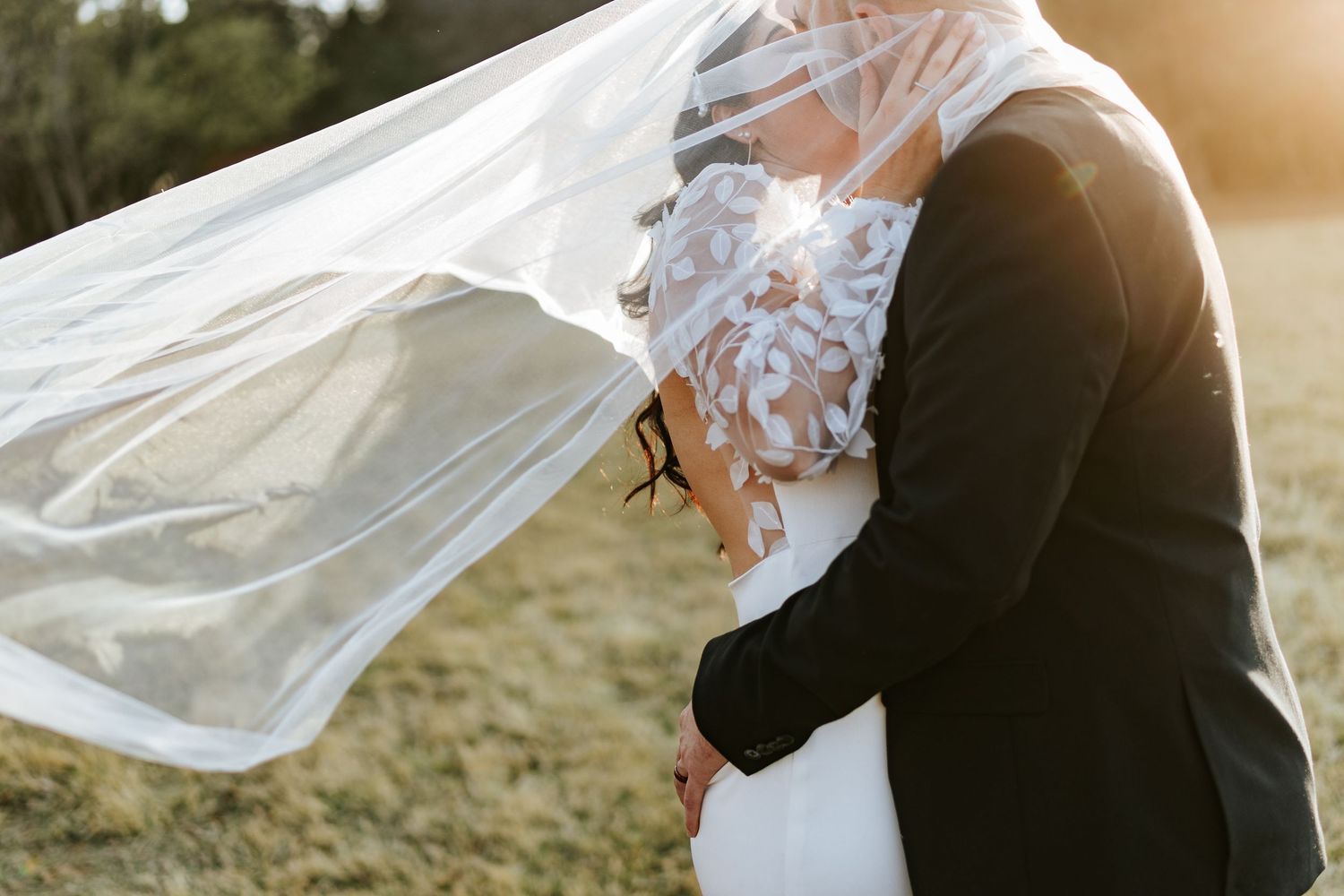 Romantic close-up of bride and groom embracing as flowing lace veil billows in the wind at sunset photographed by Laughing Lamp Photo & Film