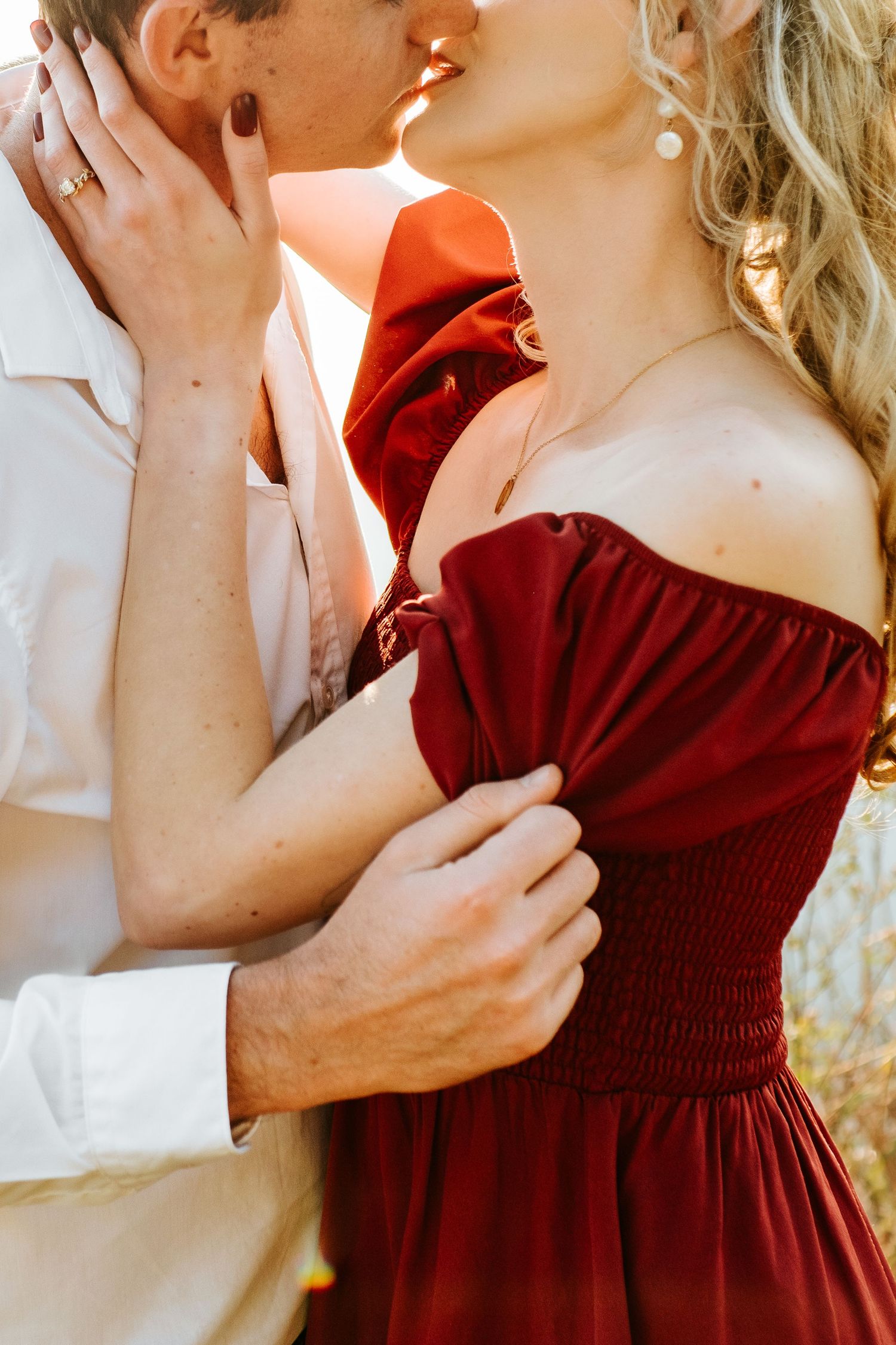 Couple sharing a romantic kiss as woman in elegant off-shoulder burgundy dress embraces man in white shirt.