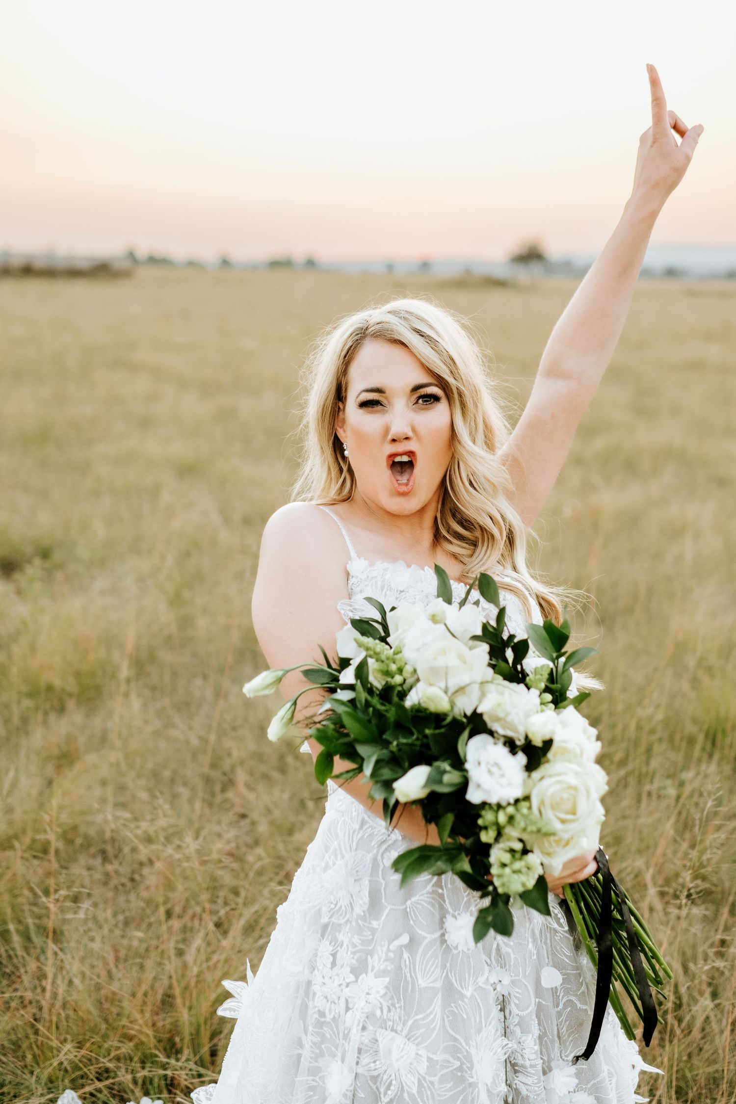 Excited blonde bride in lace gown raises hand while holding white floral bouquet in field.