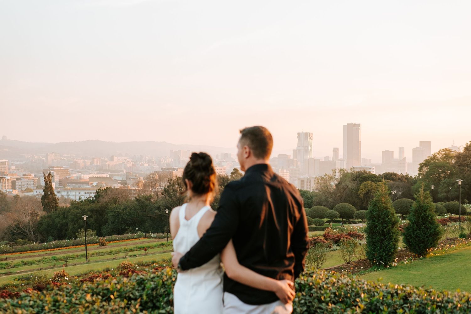Couple embraces while overlooking a city skyline surrounded by lush gardens at sunset.