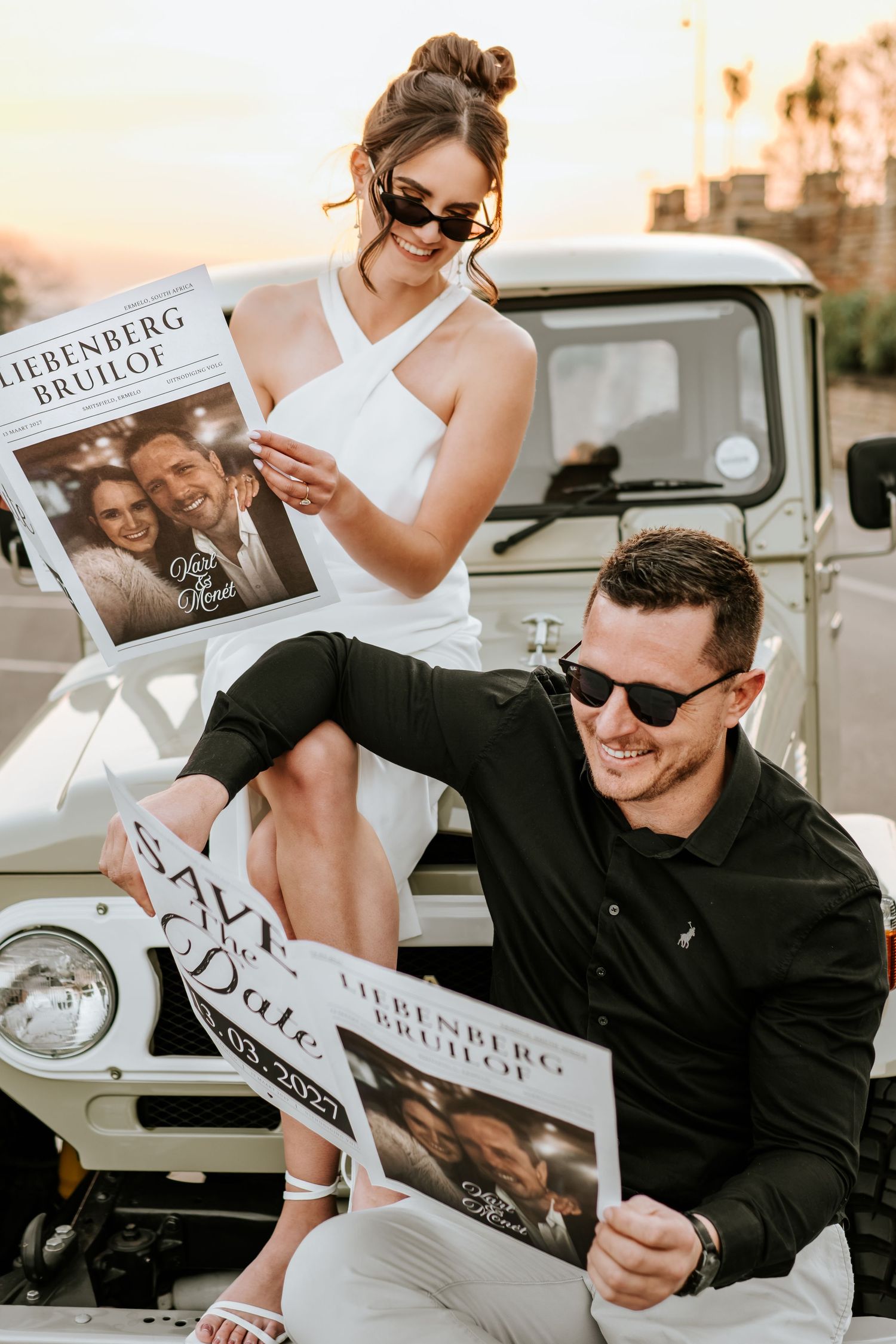 Stylish couple in sunglasses pose with wedding newspaper save-the-date cards beside a vintage white truck at sunset.