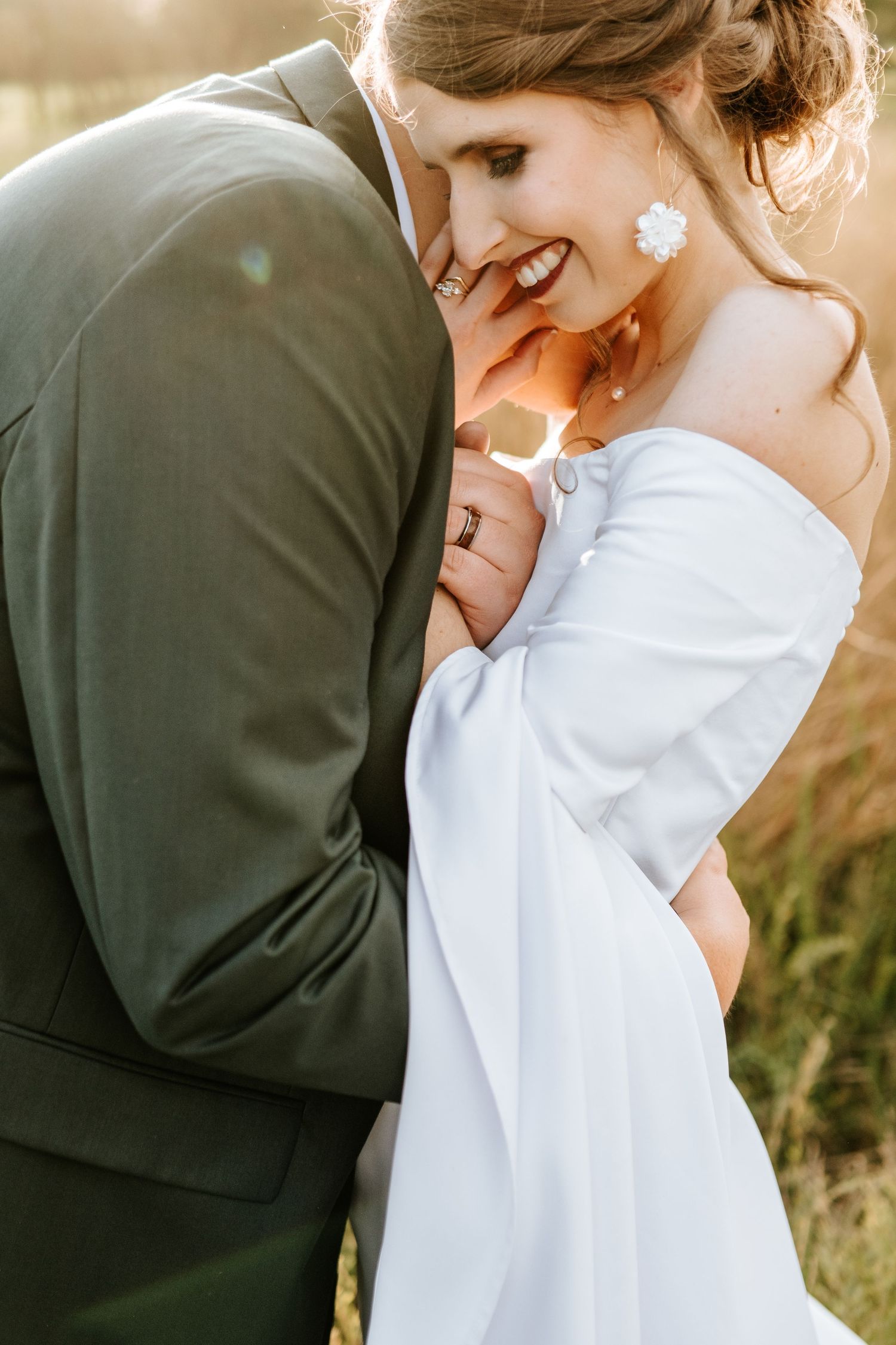 Smiling bride in off-shoulder white dress embraced by groom in green suit during golden hour.
