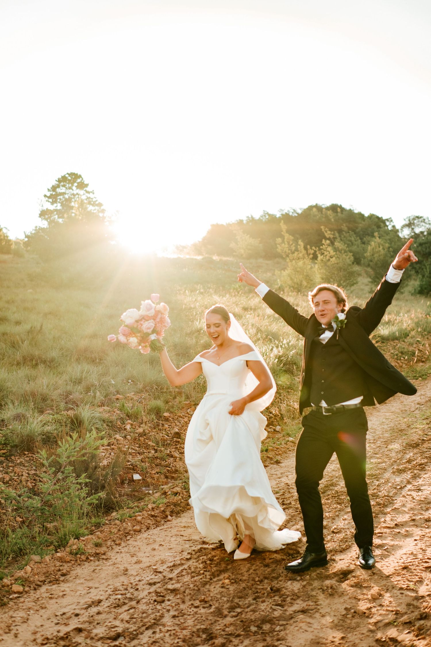 Joyful newlyweds celebrate on a dirt path at sunset, bride holding pink balloons as groom raises arms triumphantly.
