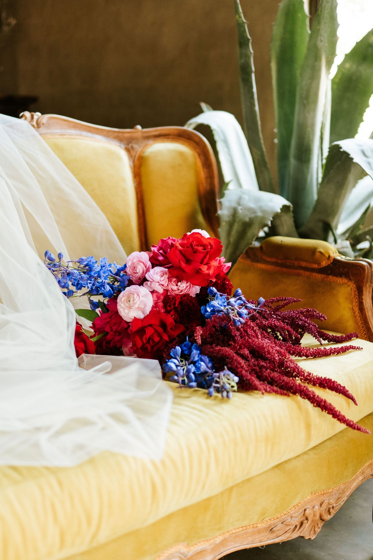 Vibrant wedding bouquet with red, pink, and blue flowers resting on vintage yellow velvet sofa with veil.