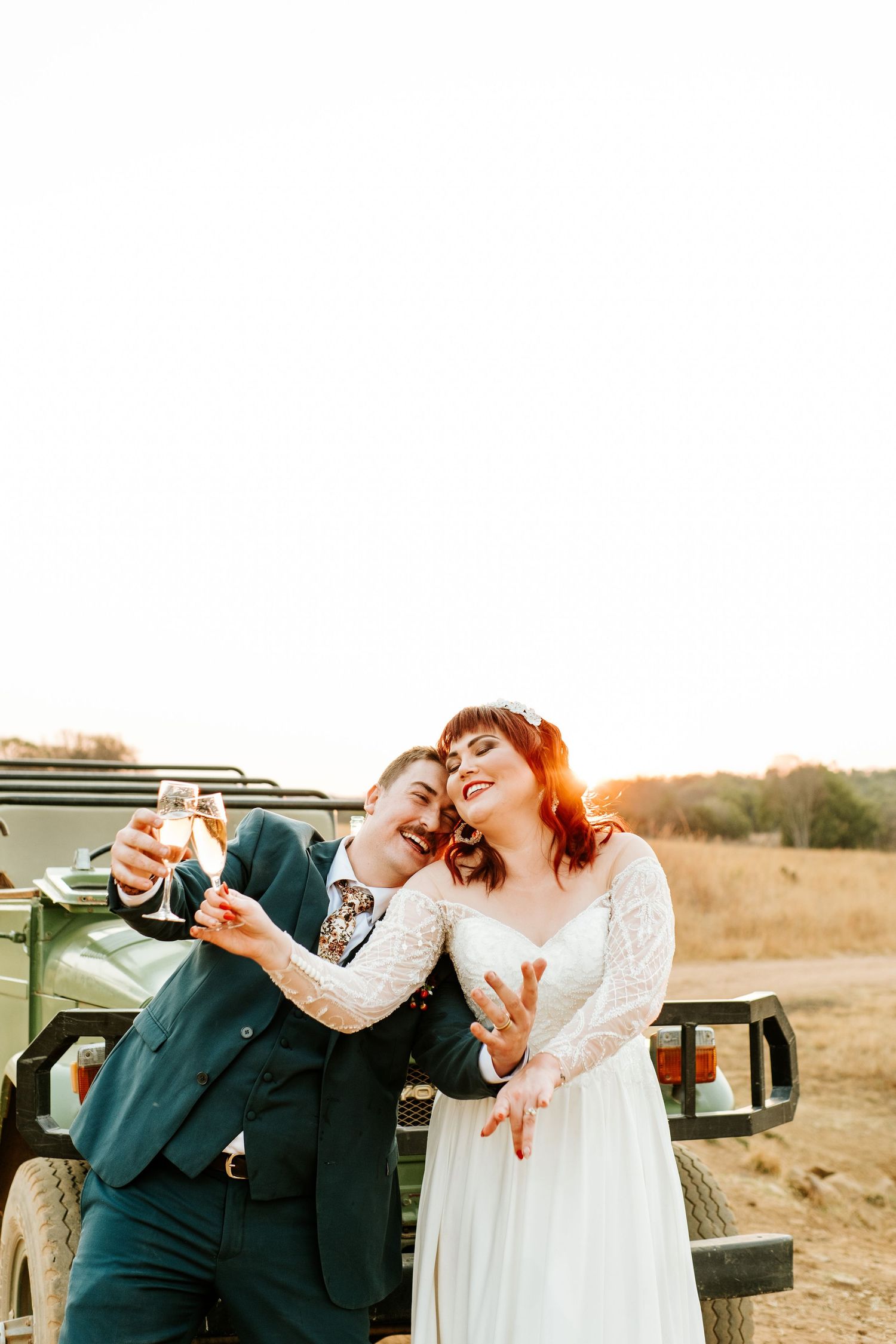 Joyful newlyweds toast champagne beside a vintage green jeep at golden hour outdoors.