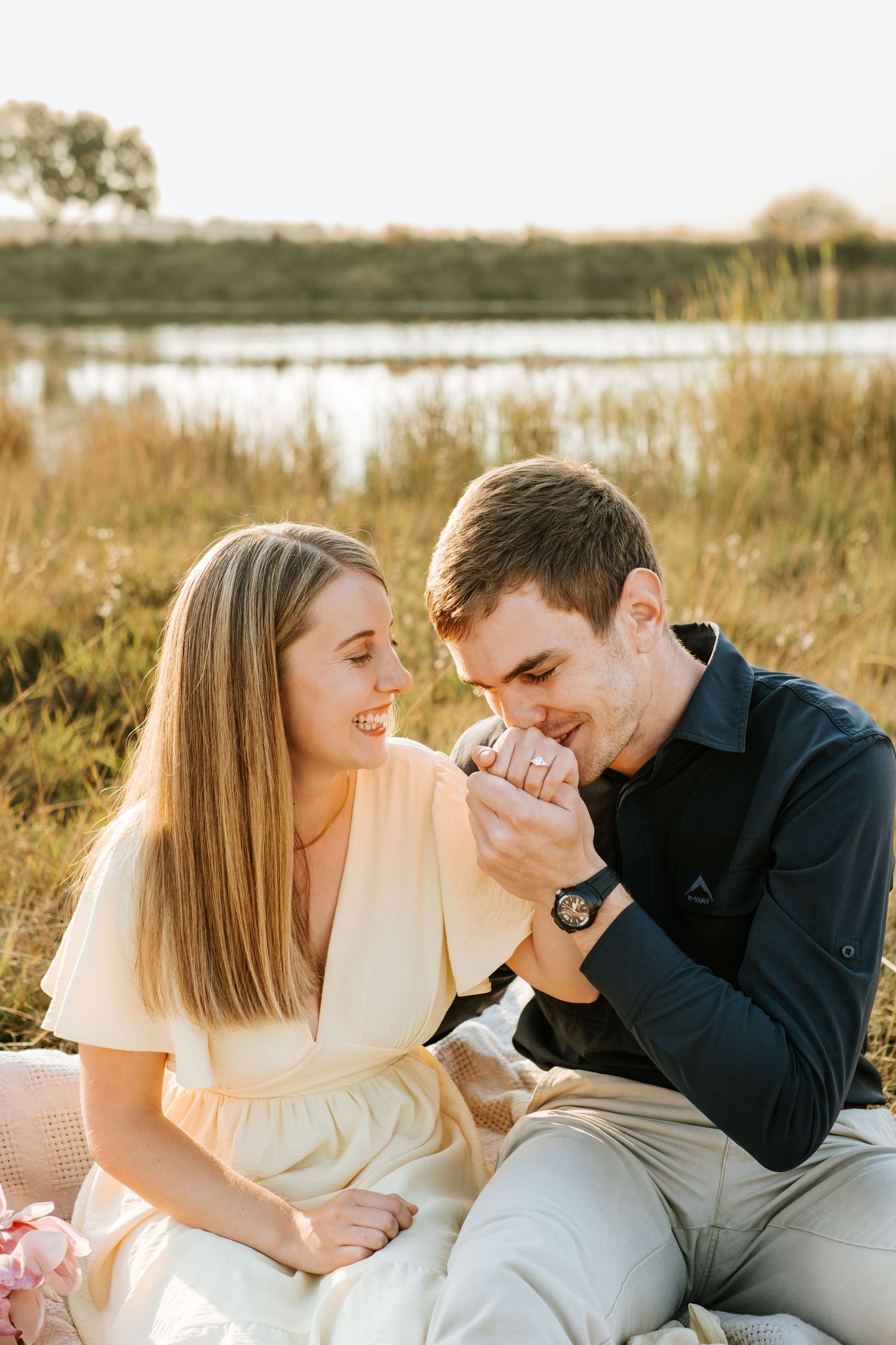 Smiling couple sitting on picnic blanket near lake as man kisses womans hand in warm golden sunlight.