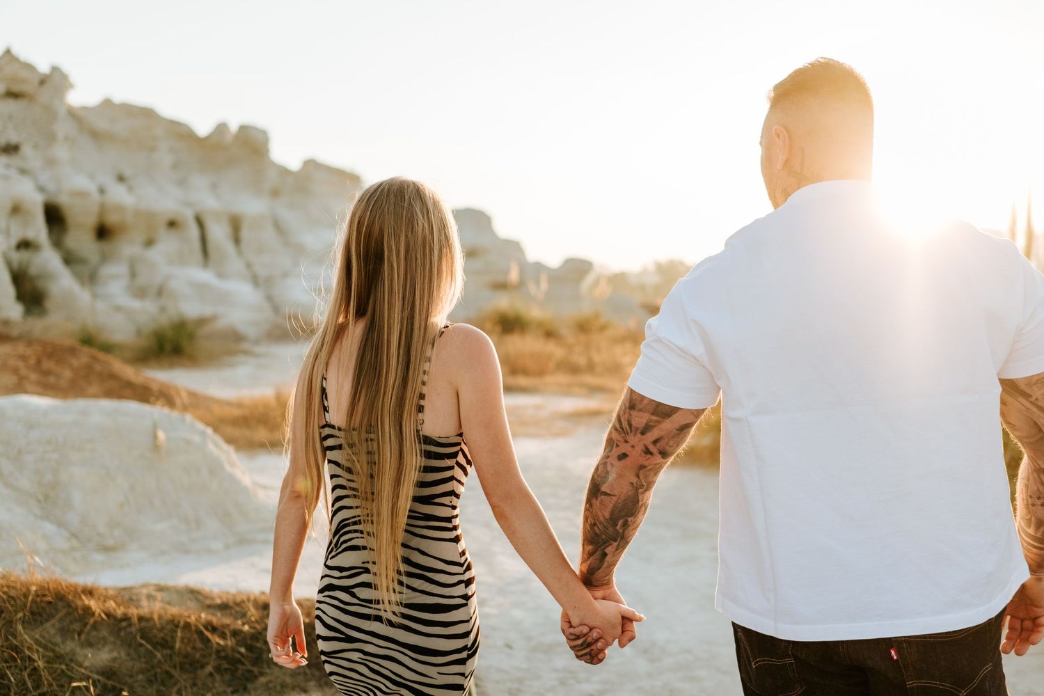 Couple holding hands walking toward white rock formations in warm golden sunset light.