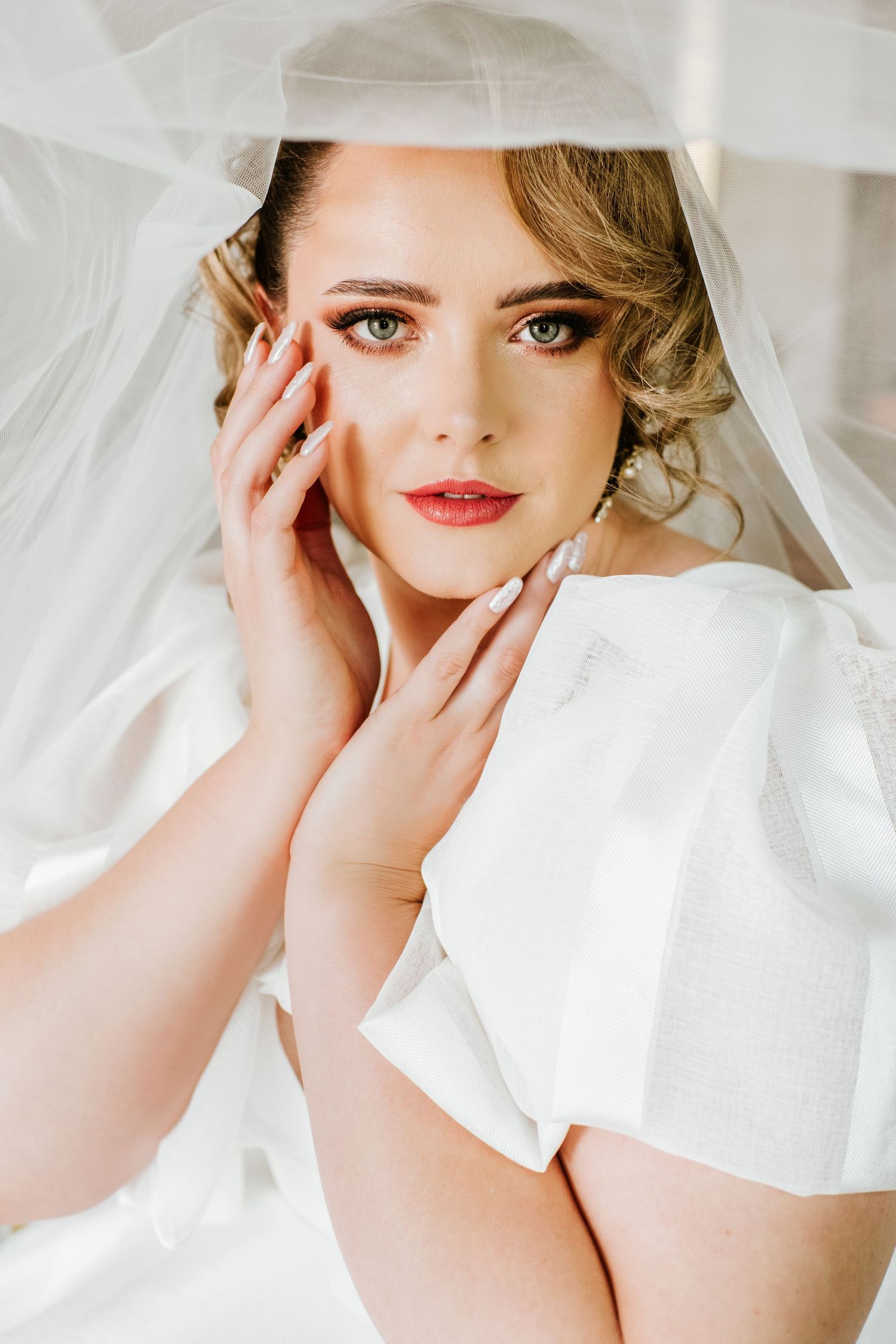 Elegant bride wearing a white veil and dress, posing with hands near face showing flawless bridal makeup.