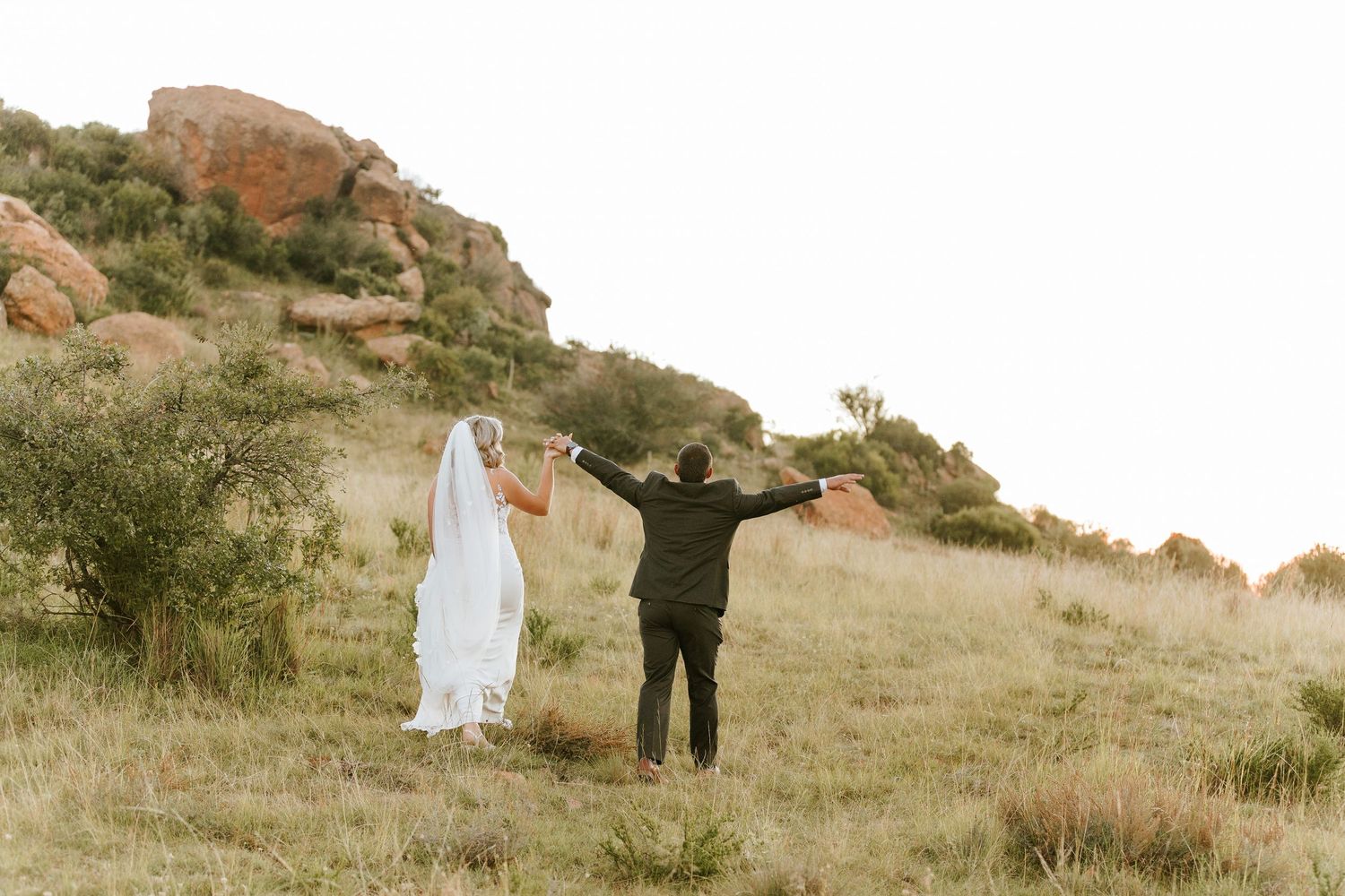 Bride and groom walk up a grassy hill with arms raised near scenic red rock formations.