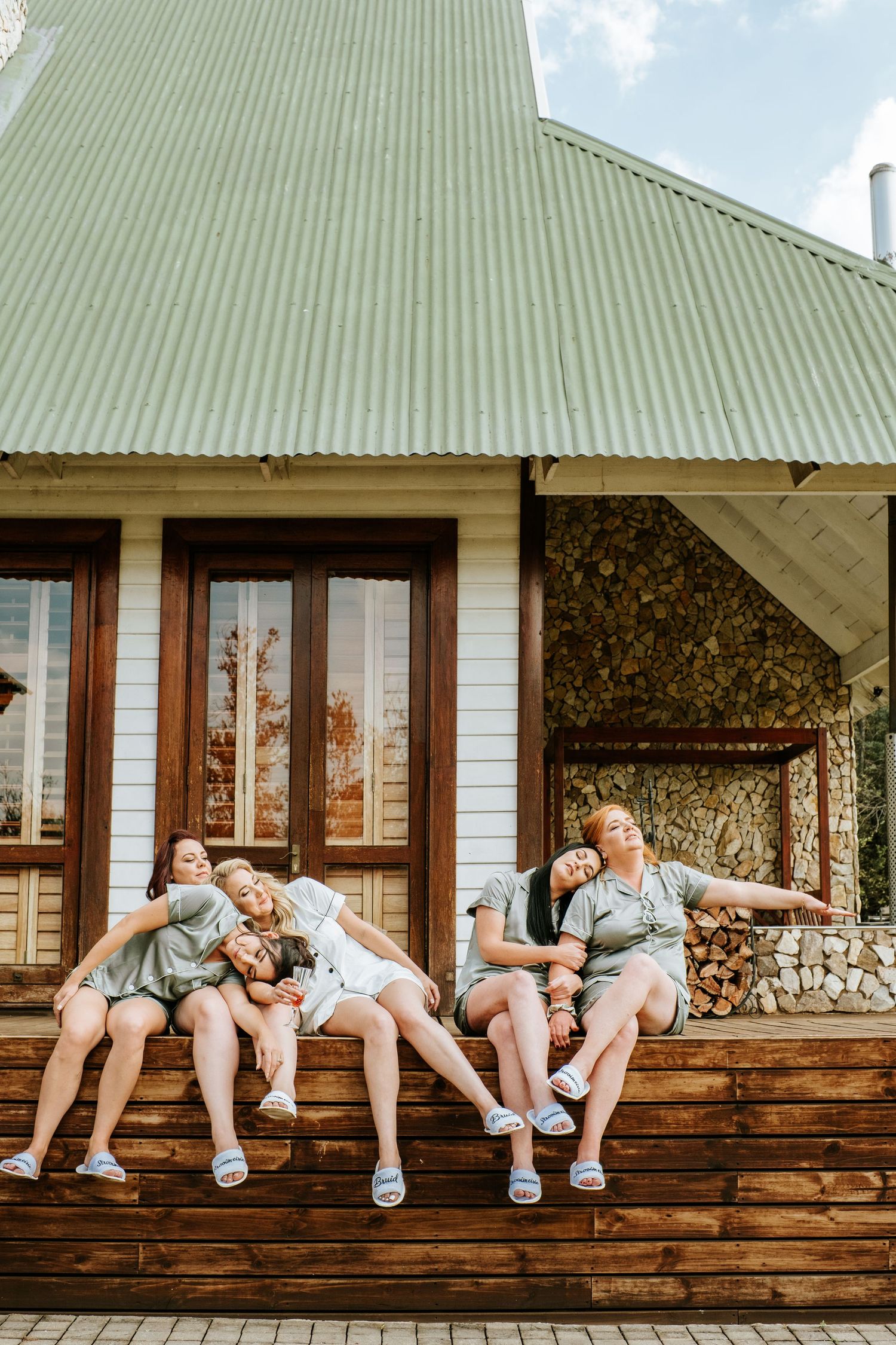 Four bridesmaids in sage green robes relaxing together on wooden deck outside rustic venue.
