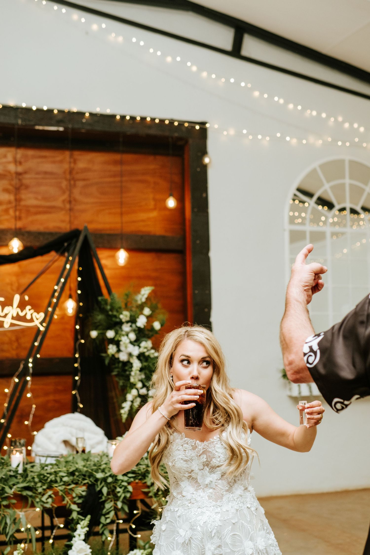 Blonde bride in lace gown drinking from a glass on dance floor with string lights in background.