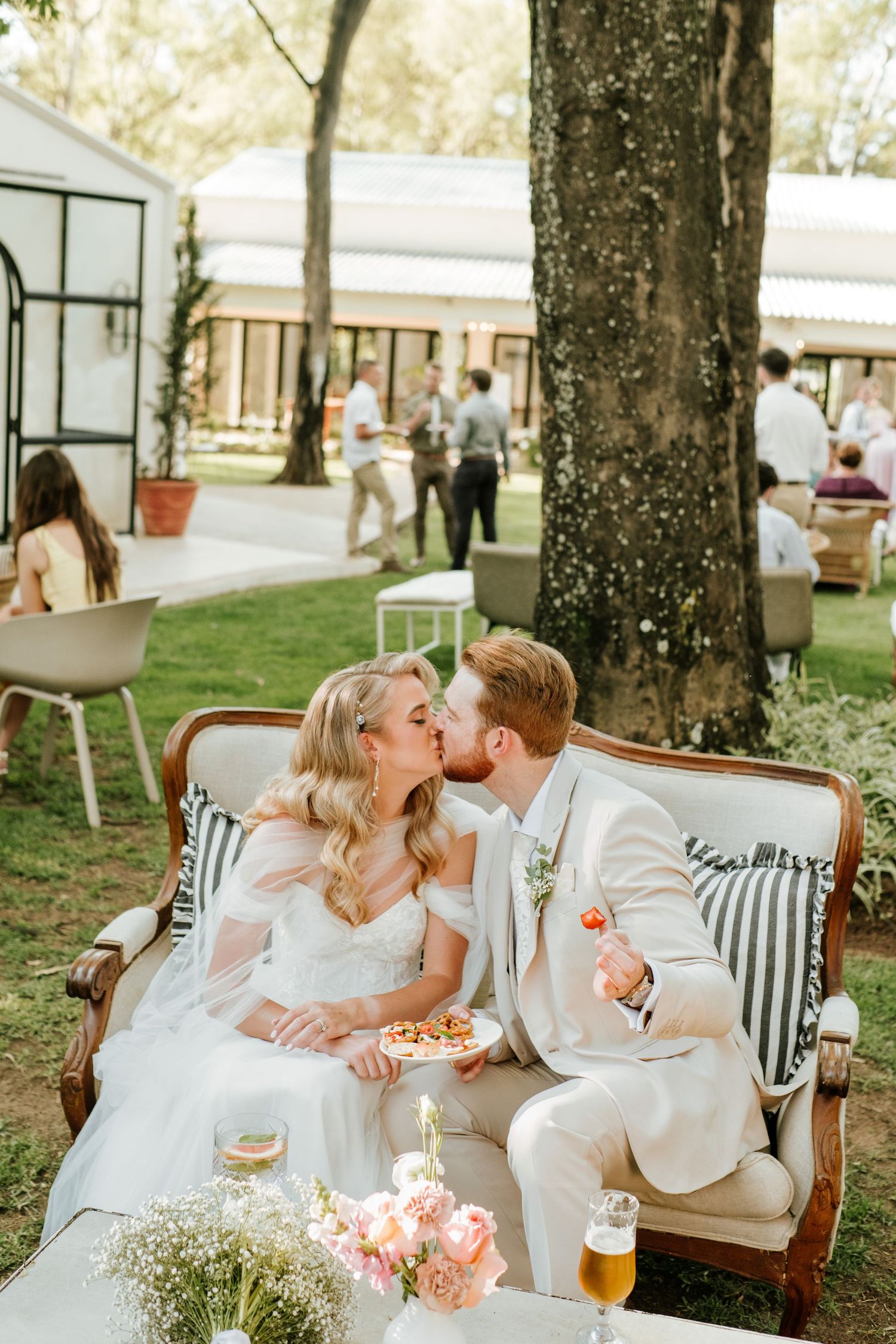 Newlyweds share a kiss on vintage sofa during outdoor garden wedding reception with guests mingling behind.