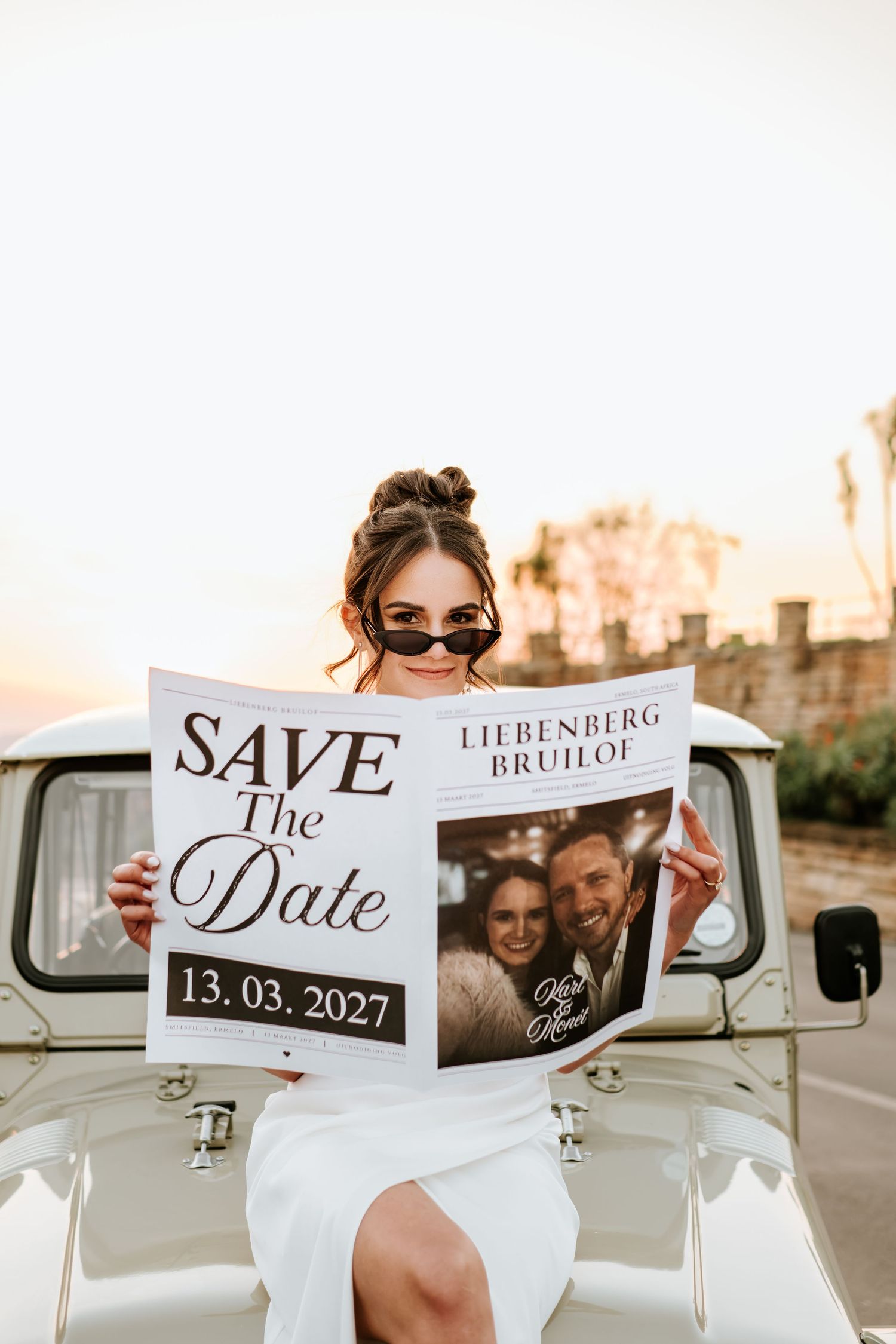 Bride holding Save the Date card for Liebenberg wedding on 13.03.2027 leaning against vintage white Toyota truck.
