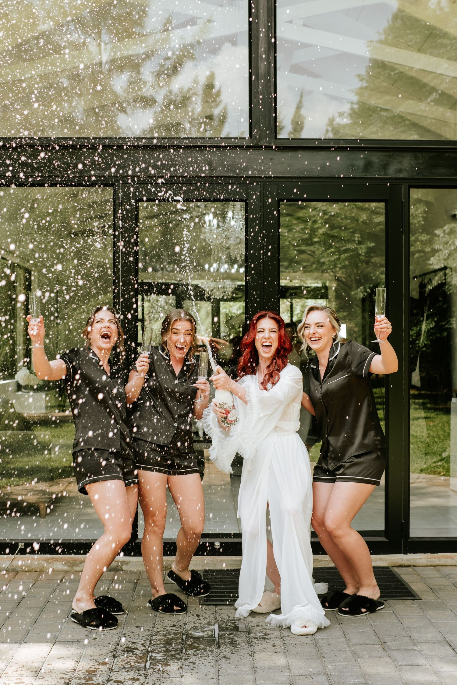 Excited bride in white robe celebrating with bridesmaids in black pajamas under falling confetti.