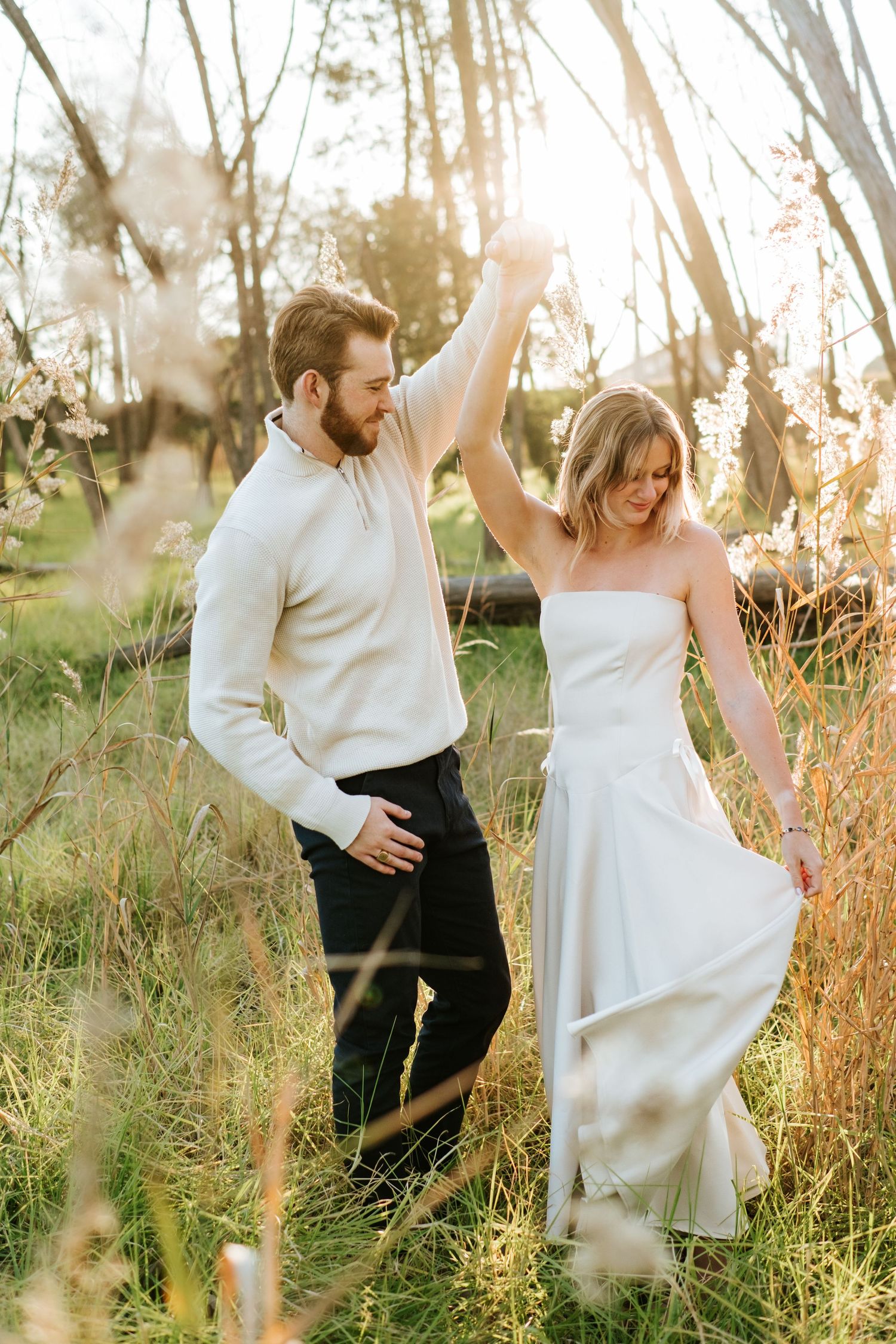Bride in white gown being twirled by groom in white sweater through tall grass at sunset.