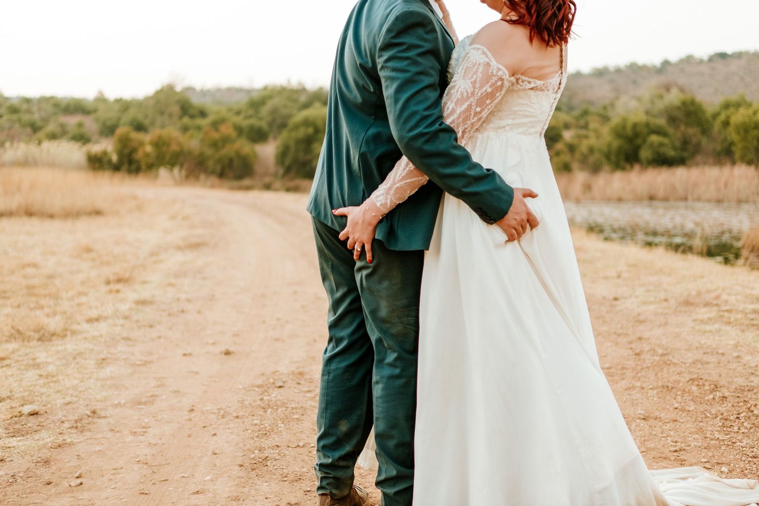 Bride and groom embracing on a rustic dirt path surrounded by golden fields on their wedding day.