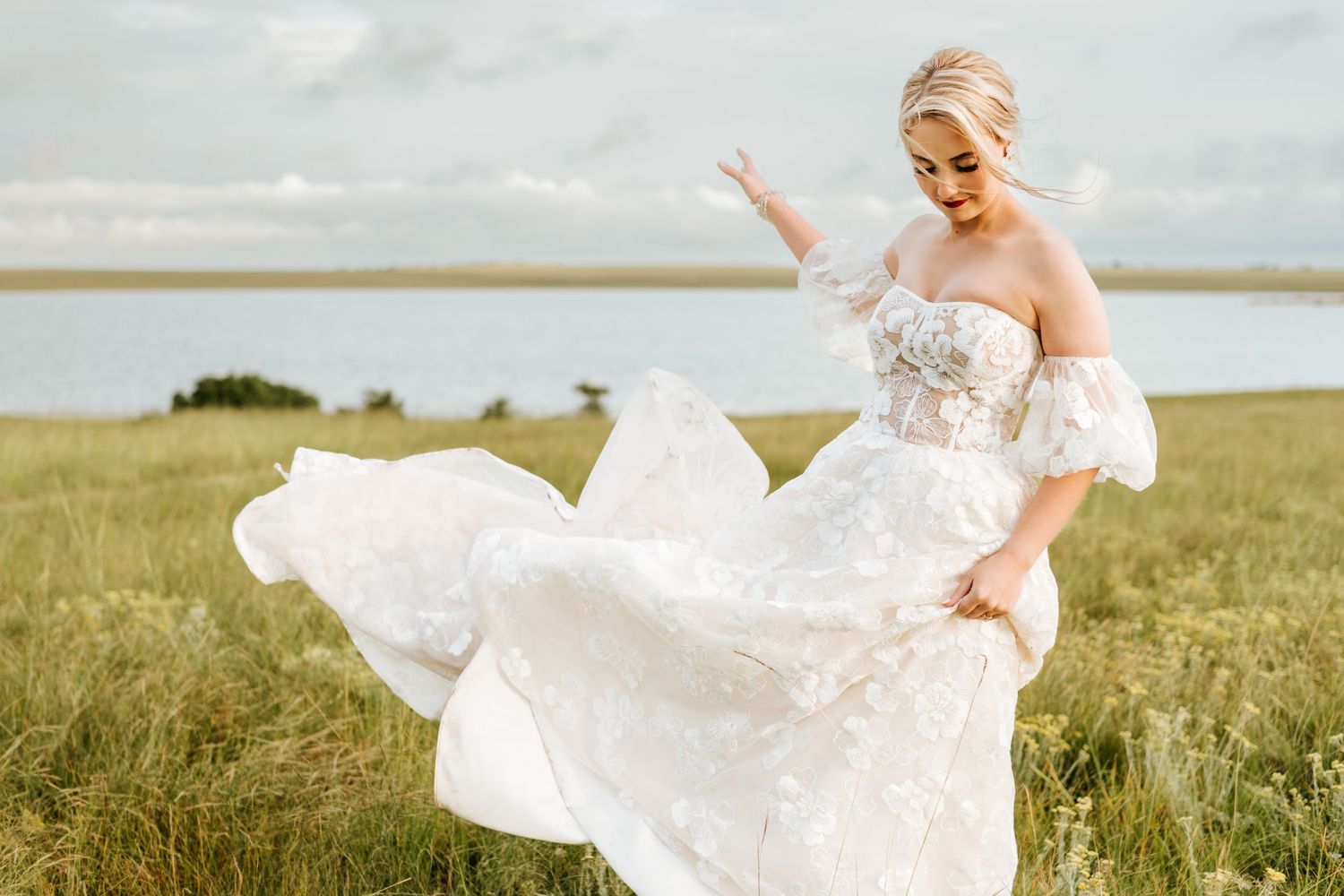 Bride in floral off-shoulder gown twirls dress in green meadow with lake backdrop.
