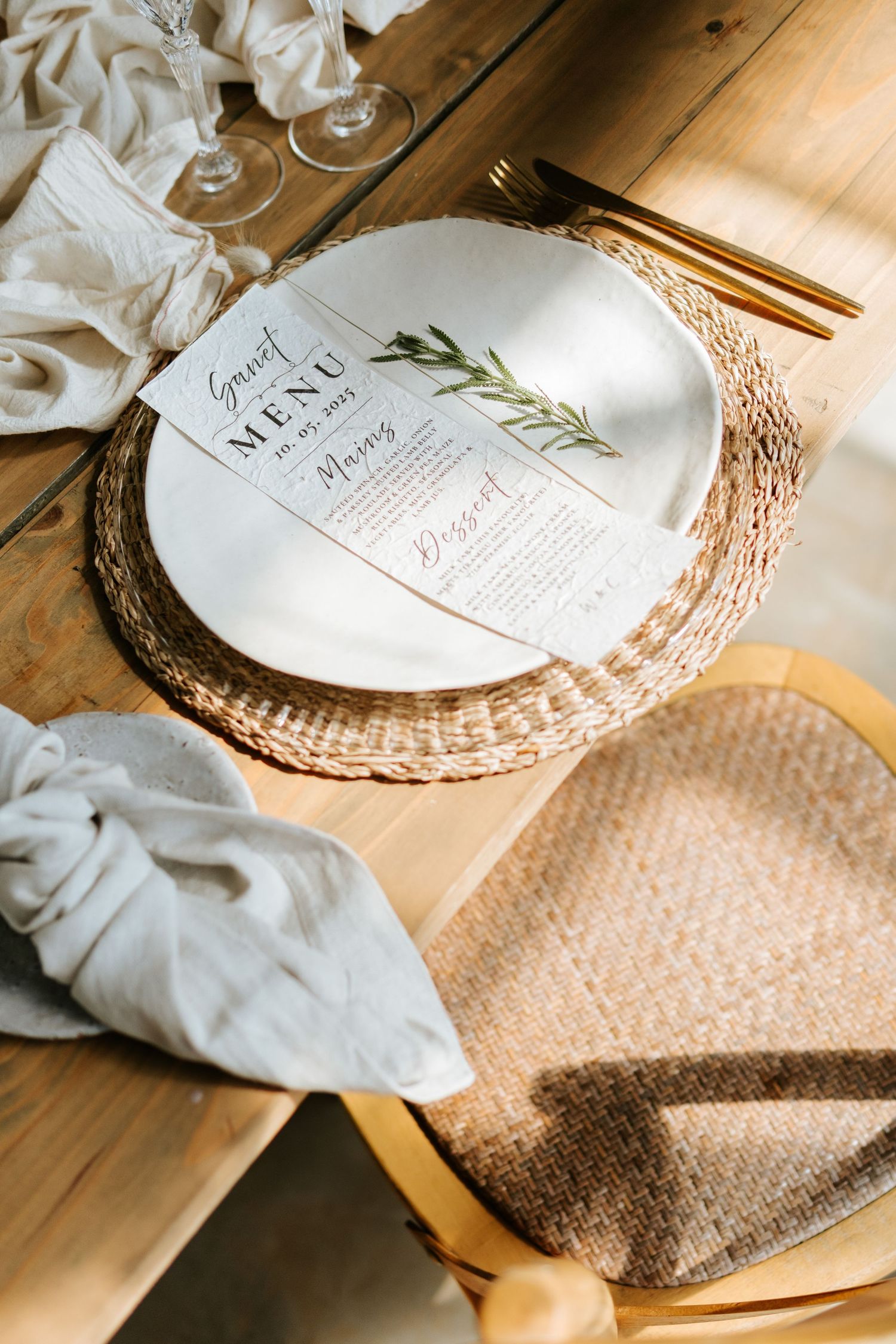 Elegant wedding place setting with woven charger, white plate, guest menu card, and gold cutlery on wood.