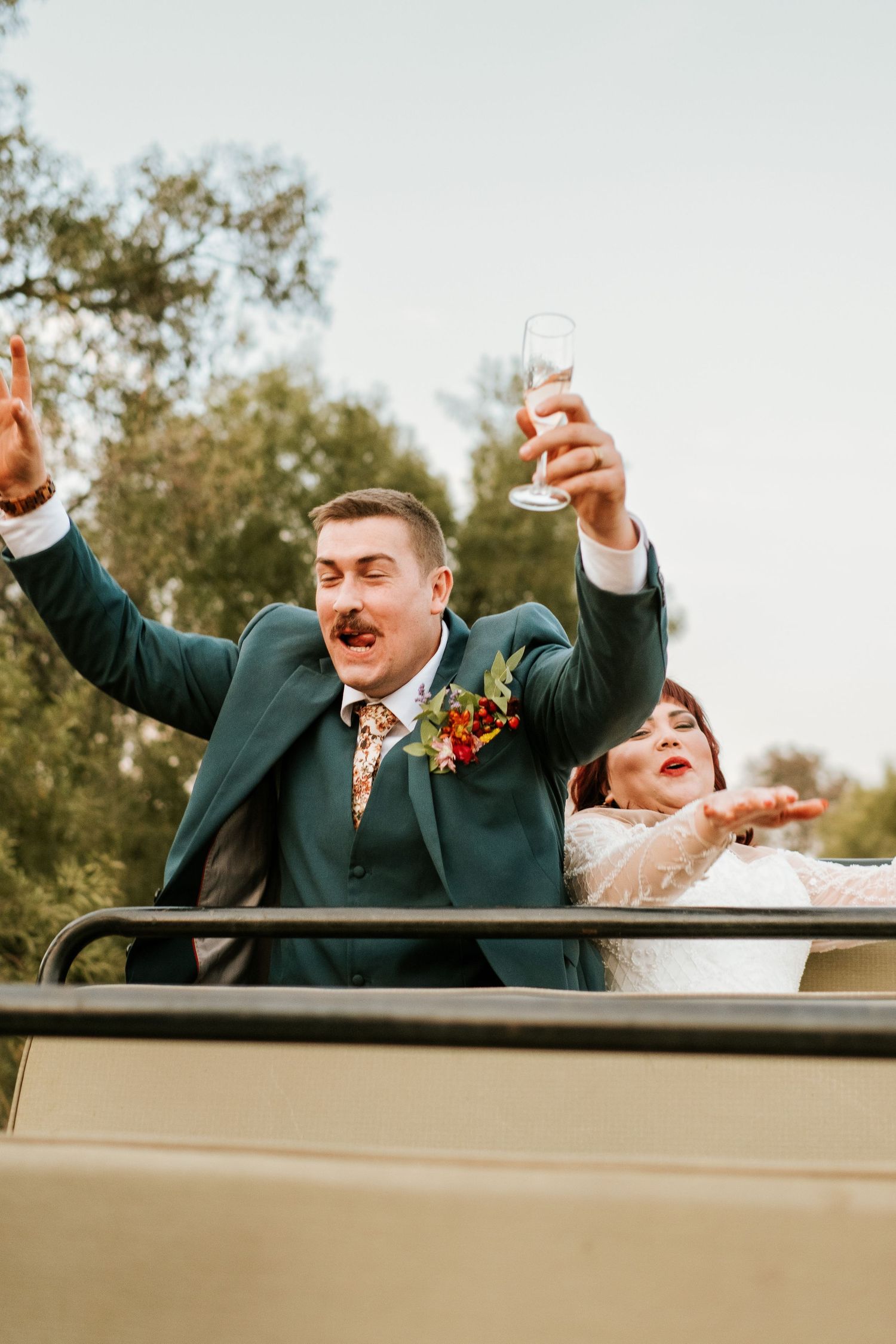 Joyful groom in green suit raises champagne glass while bride celebrates in safari vehicle photographed by Laughing Lamp Photo & Film