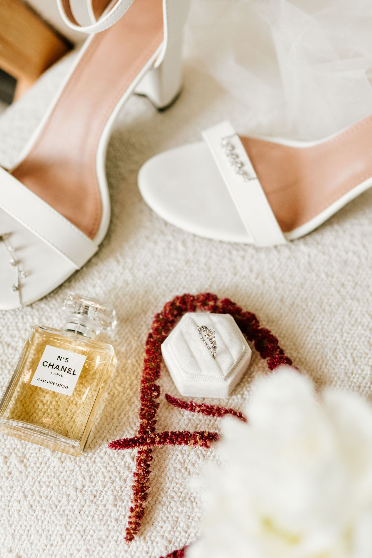 Wedding flat lay featuring Chanel No.5 perfume, white heeled bridal shoes, ring box, and red berry garland.