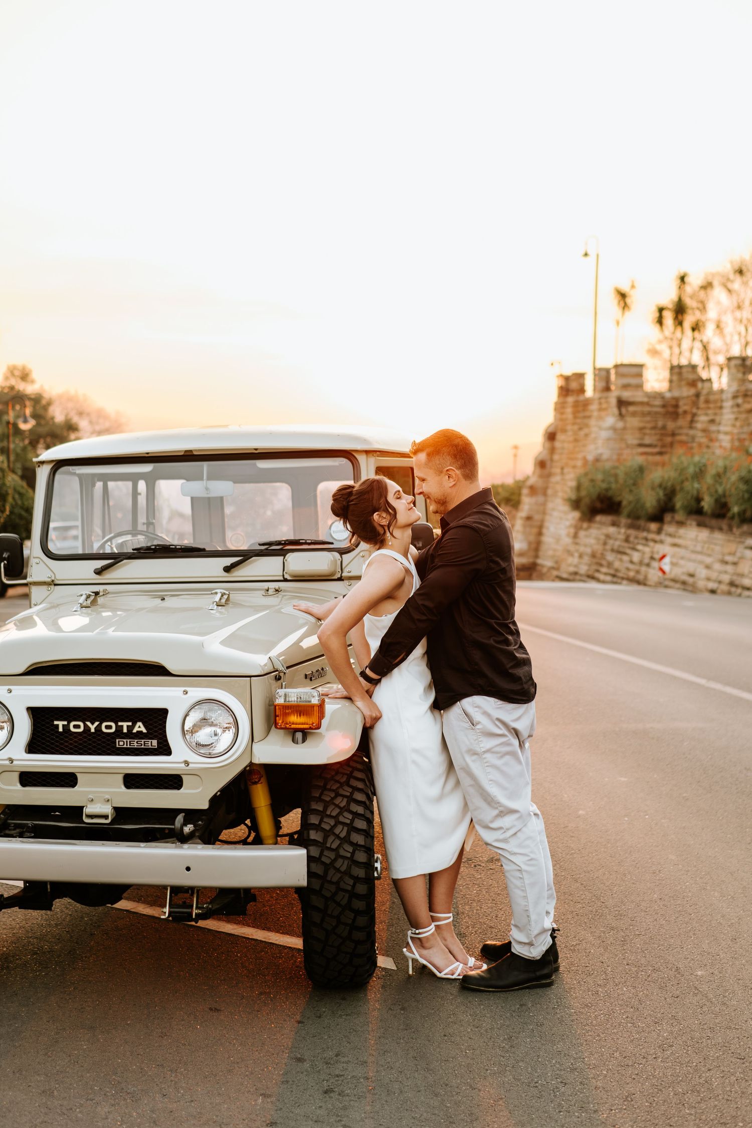 Couple shares a tender kiss leaning against a vintage Toyota FJ40 at a golden sunset.