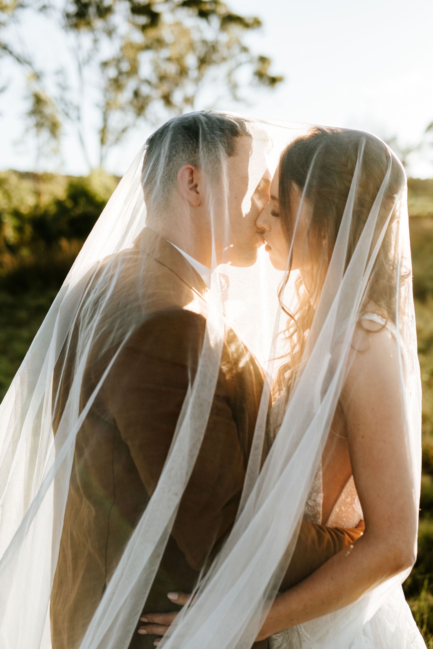 Romantic bride and groom nearly kissing under flowing white veil with warm golden backlight in outdoor setting.