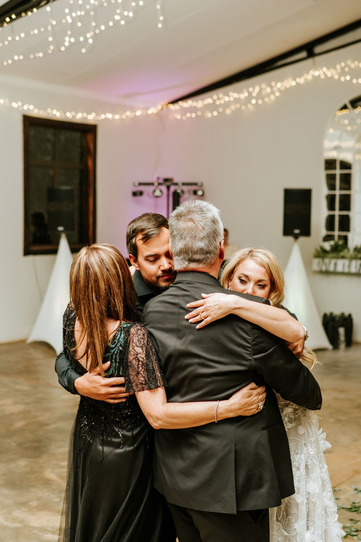Four family members sharing a group hug on dance floor under fairy lights at wedding reception.