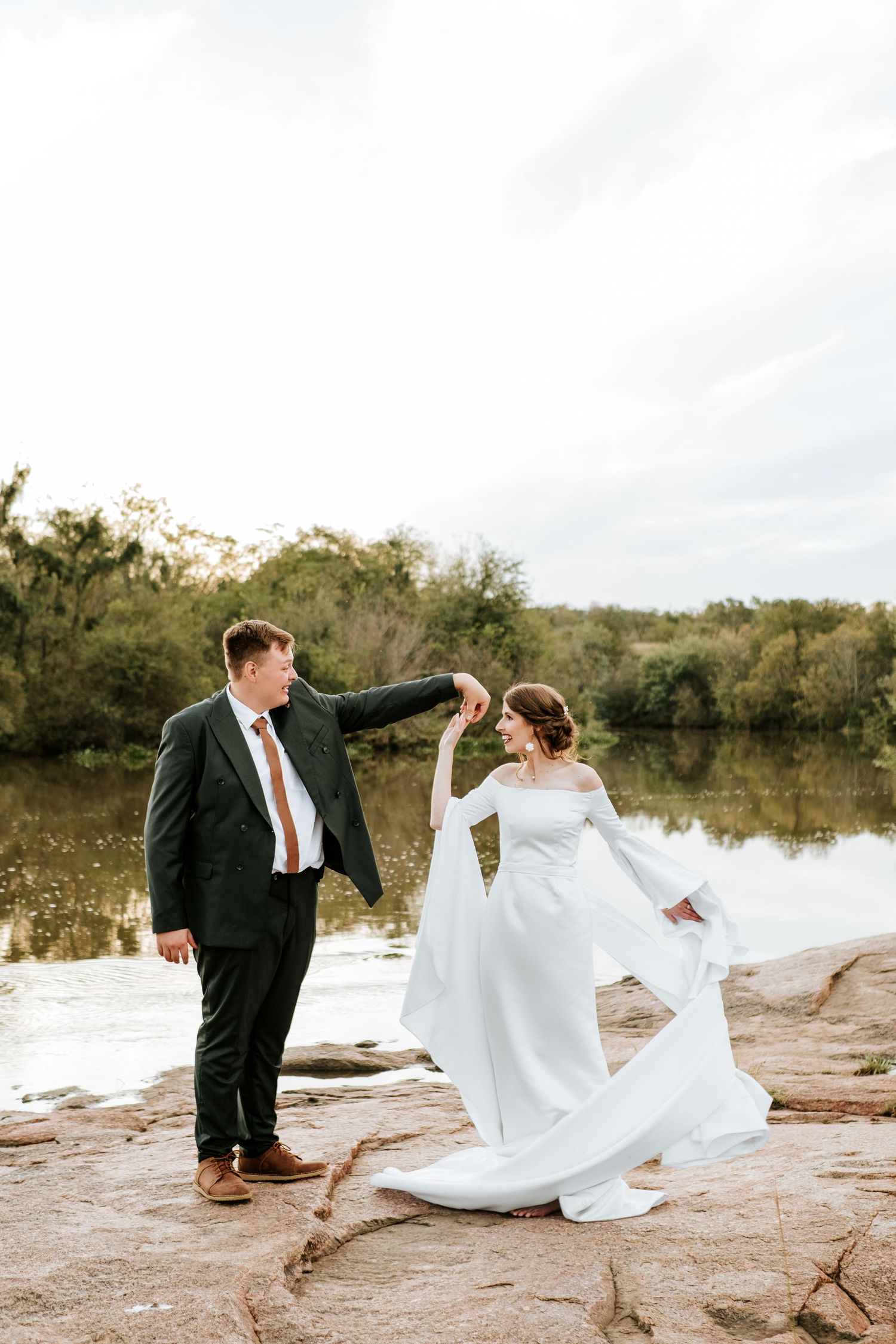 Bride in flowing white gown being twirled by groom in dark suit near scenic river backdrop.