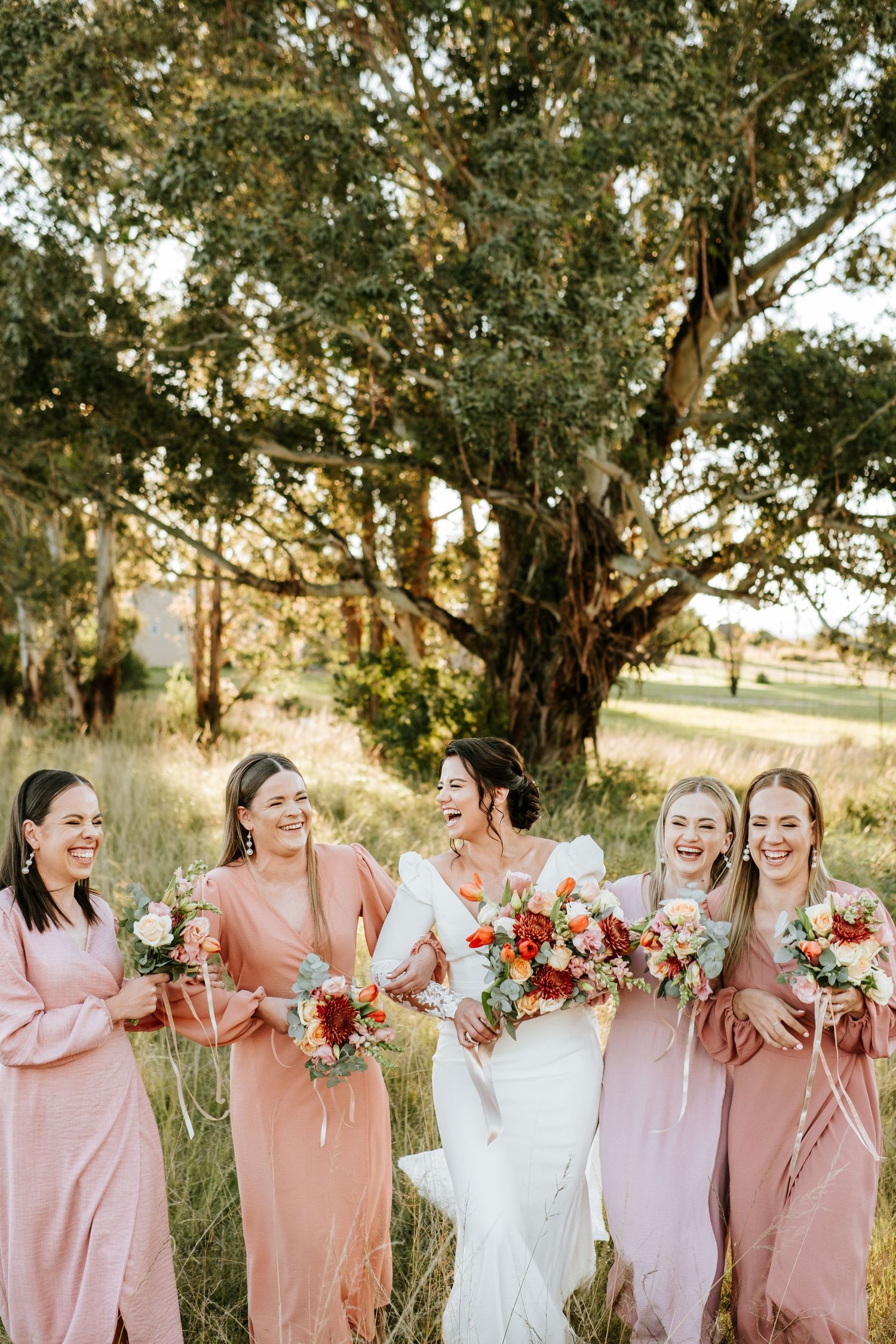 Bride laughing joyfully with four bridesmaids in pink dresses holding colorful bouquets outdoors.
