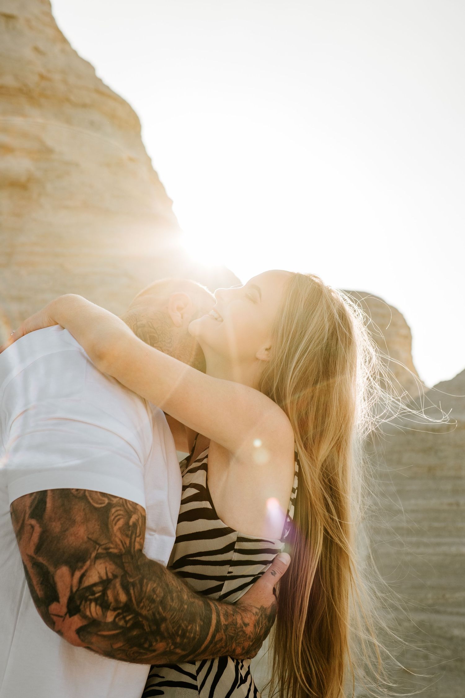 Tattooed groom kisses bride backlit by bright sunlight against sandy rock formations outdoors.