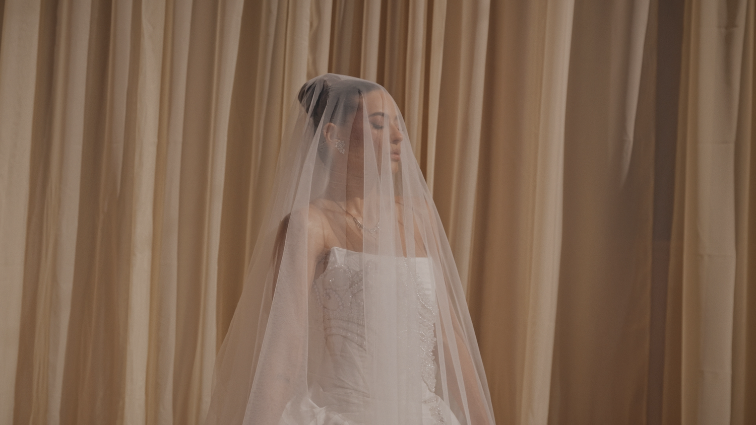 Serene bride in white strapless gown and long veil posing against elegant golden draped curtains.