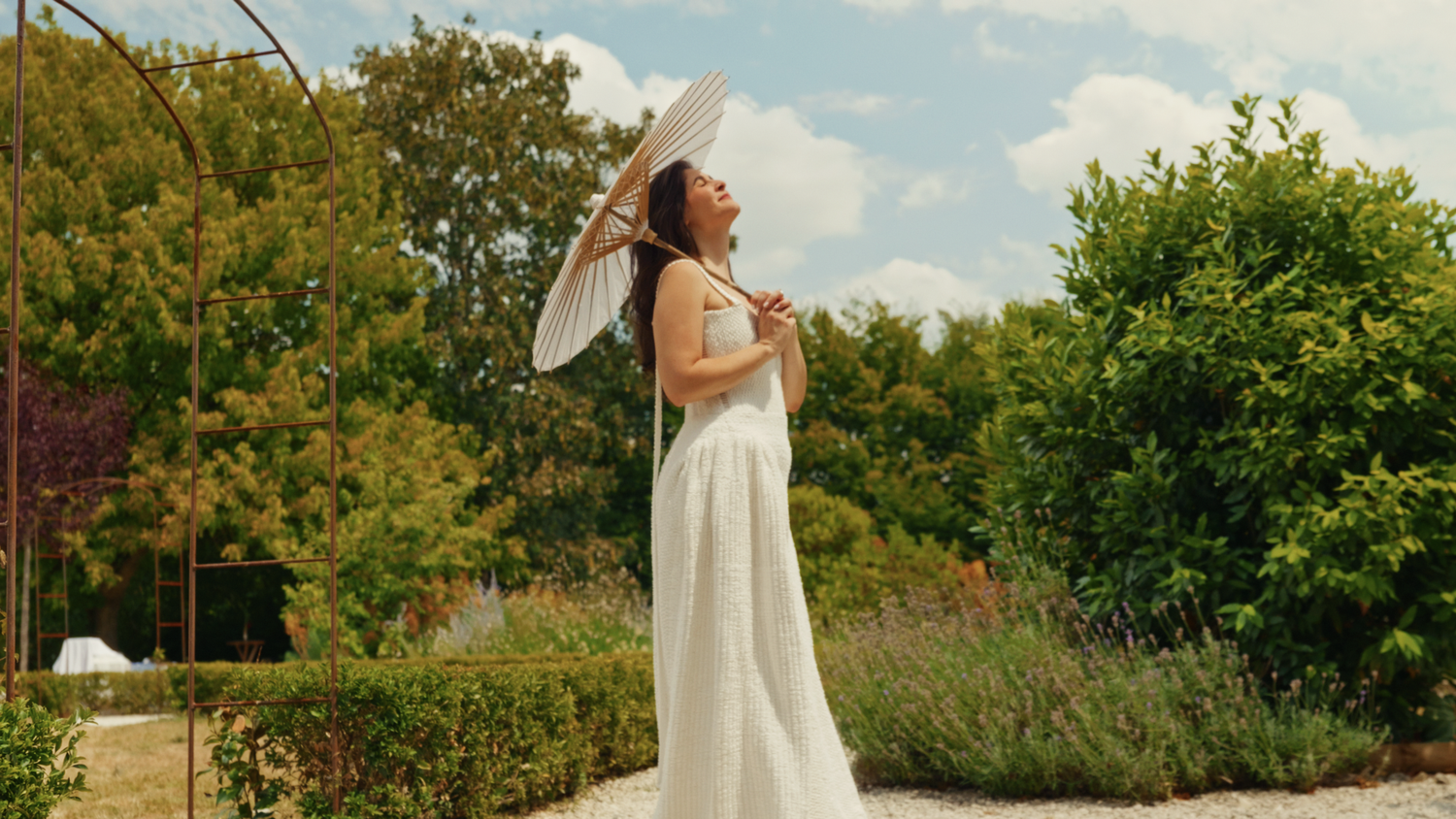 Elegant bride in a flowing white gown holding a parasol in a lush garden on a sunny day.