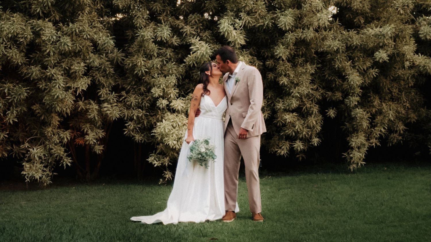 Bride and groom share romantic kiss in front of lush white flowering bushes on wedding day.
