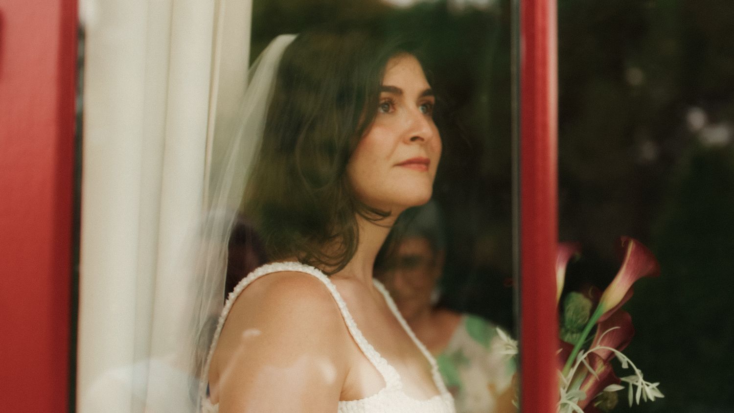 A bride in a white dress and veil gazes upward by a red door, holding a floral bouquet on her wedding day.