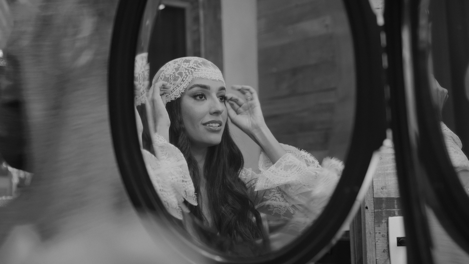 Black and white portrait of a bride adjusting her lace headpiece in a round mirror.