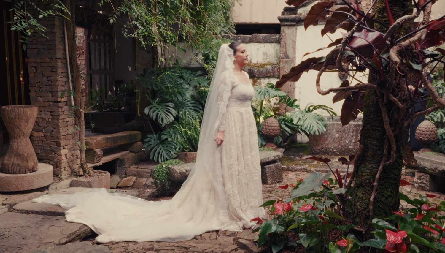 Bride in long-sleeve lace wedding gown with cathedral veil standing in a lush garden courtyard.