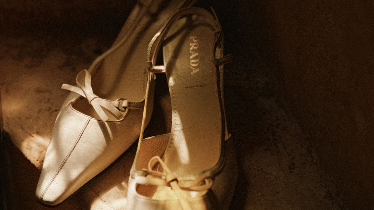 Elegant Prada slingback heels with bow detail in cream leather, displayed in warm golden light.