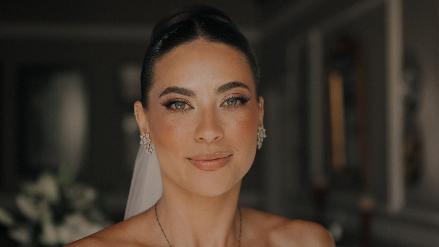 Bride with elegant updo, diamond earrings, and veil poses for a close-up wedding portrait with natural glam makeup.