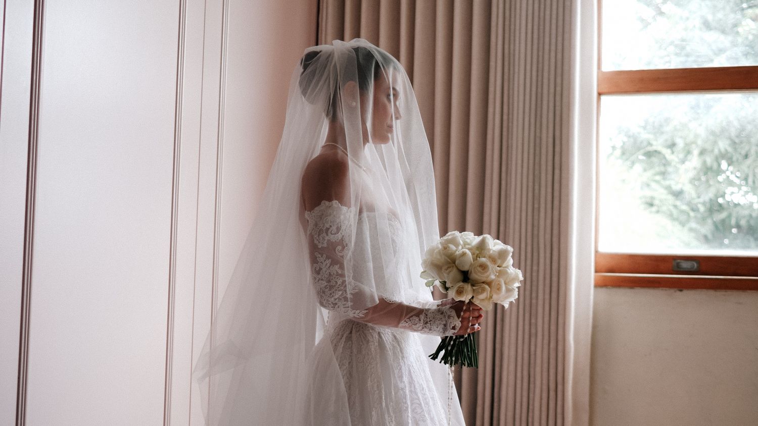 Bride in elegant lace wedding dress and veil holding white bouquet standing by window in soft natural light.