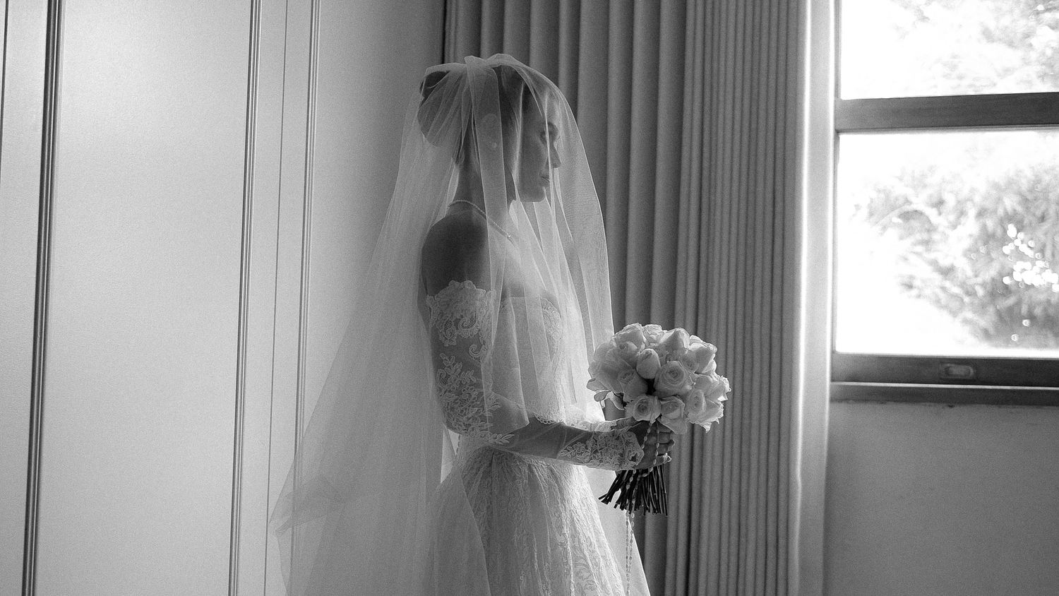 Elegant bride in lace wedding gown and veil holds white bouquet by window in black and white.