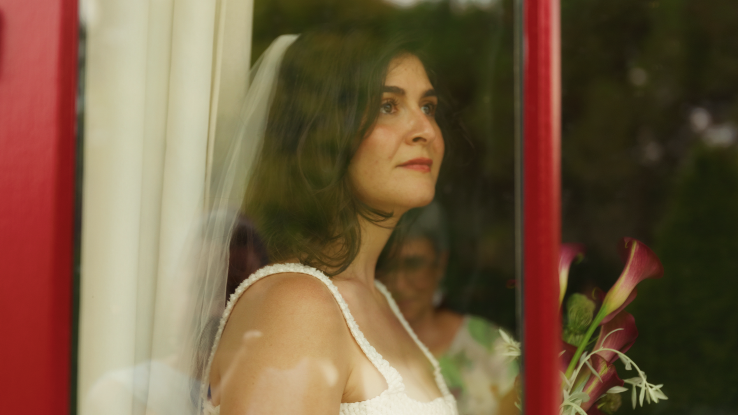 Beautiful bride in a white gown and veil gazing upward through a red-framed window.