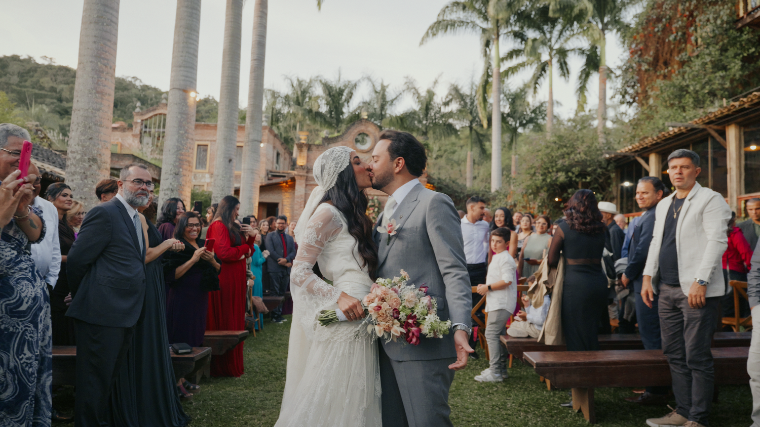 Bride and groom share first kiss at outdoor wedding ceremony surrounded by guests and palm trees.