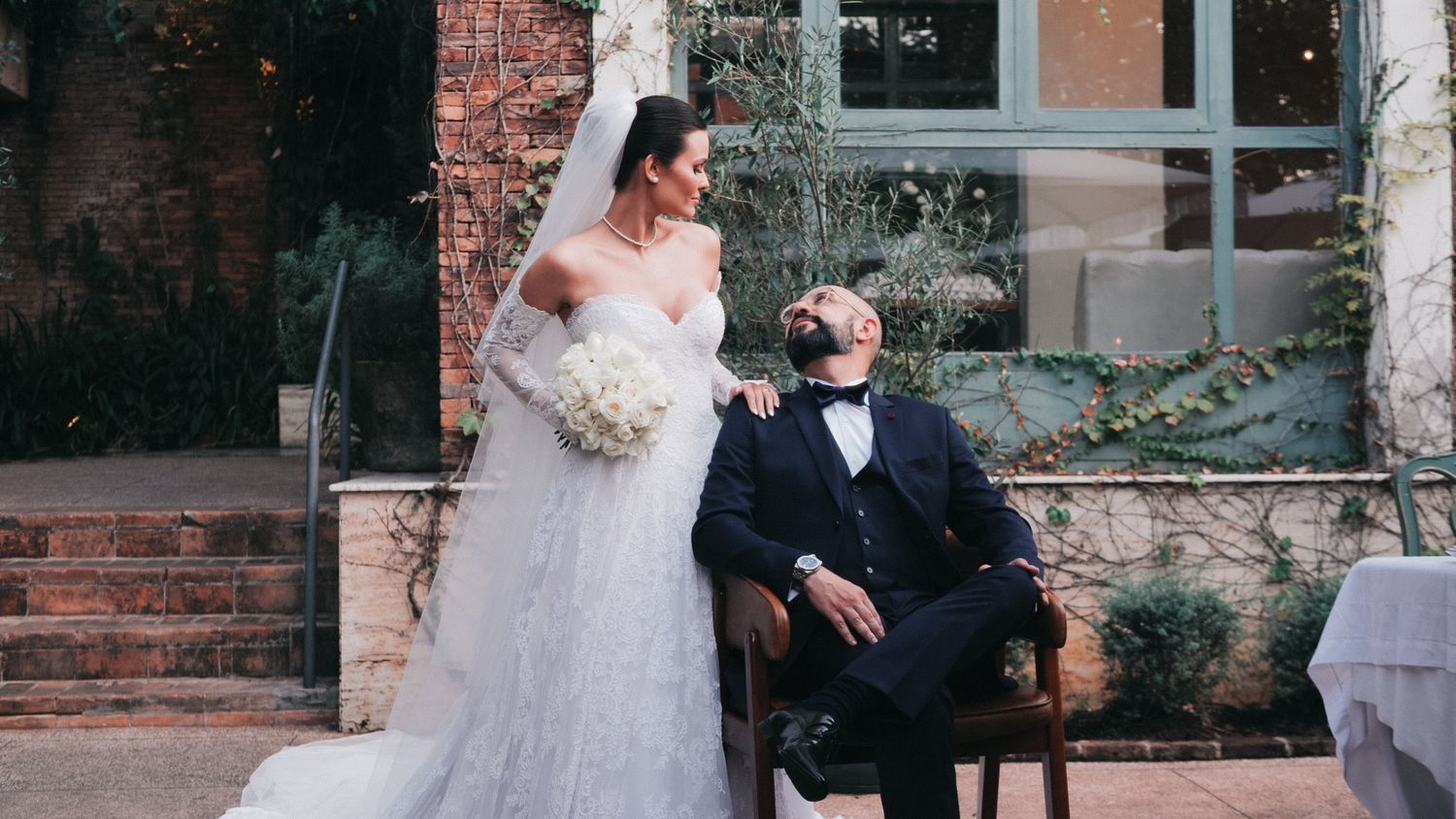 Elegant bride in white gown with veil stands beside groom in tuxedo seated outdoors at a romantic wedding venue.