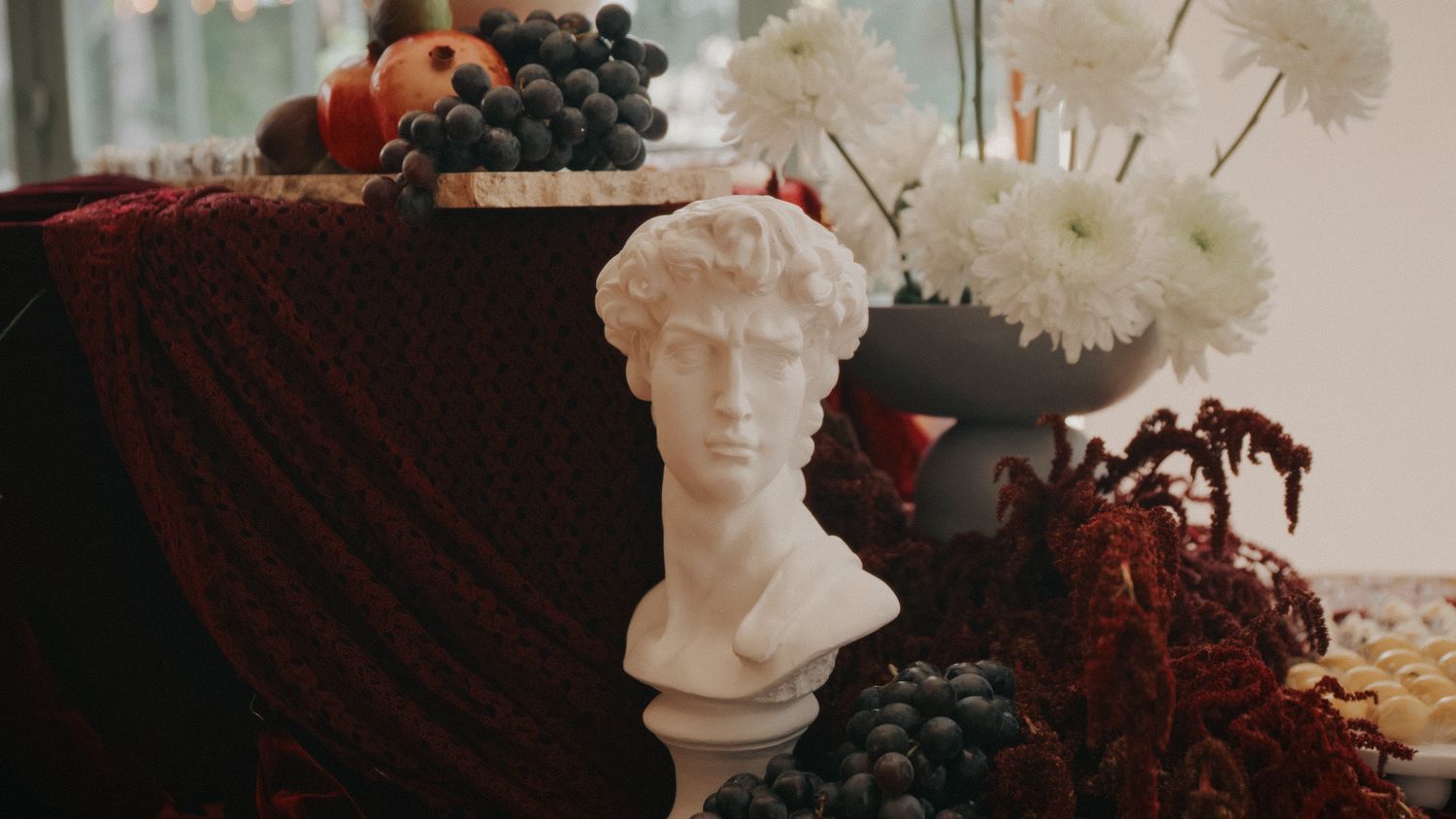 White marble David bust sculpture surrounded by grapes, white chrysanthemums, and red amaranth in an elegant still life.