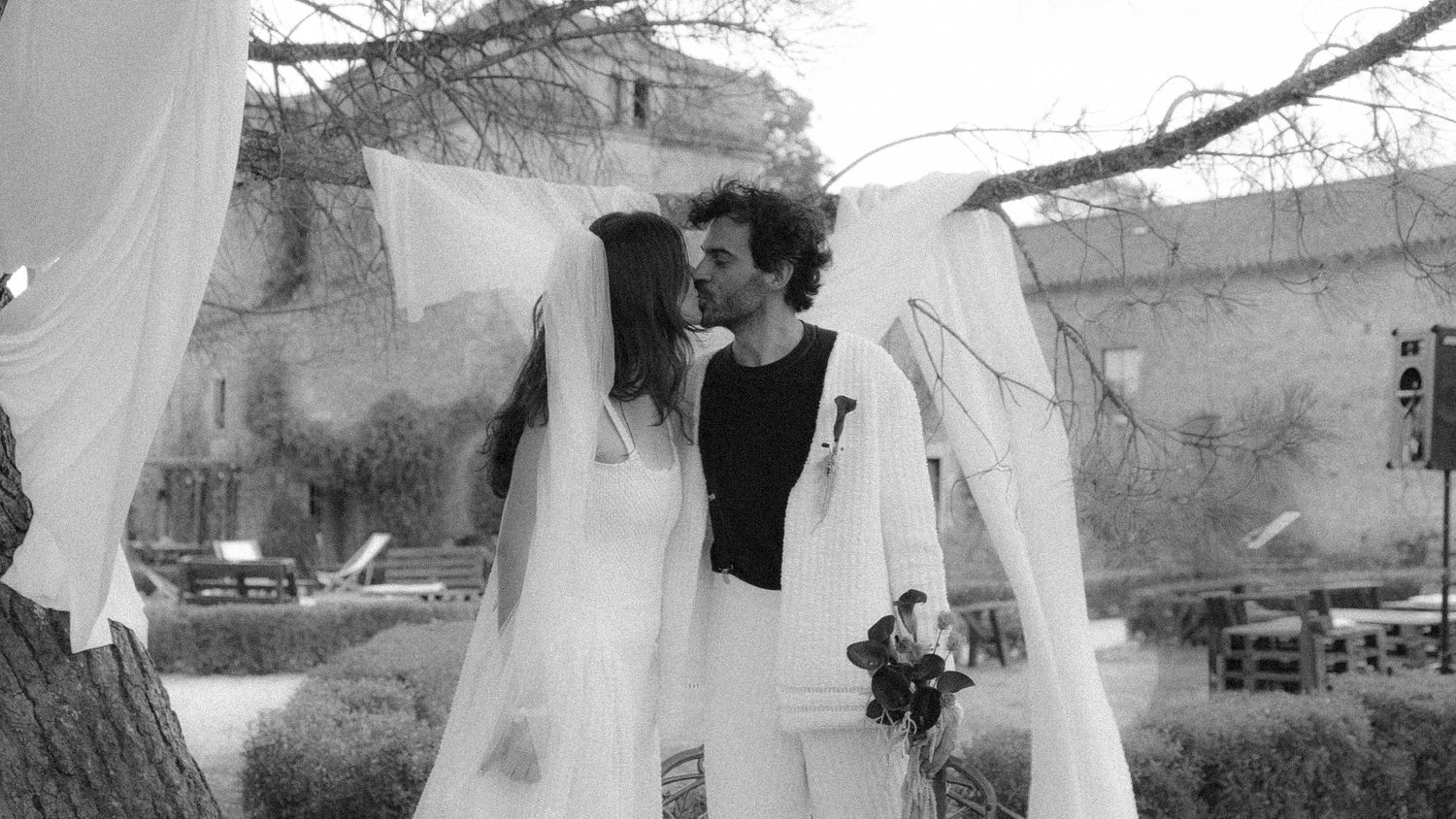 Black and white photo of a couple sharing a kiss at an outdoor wedding ceremony under a draped arch.