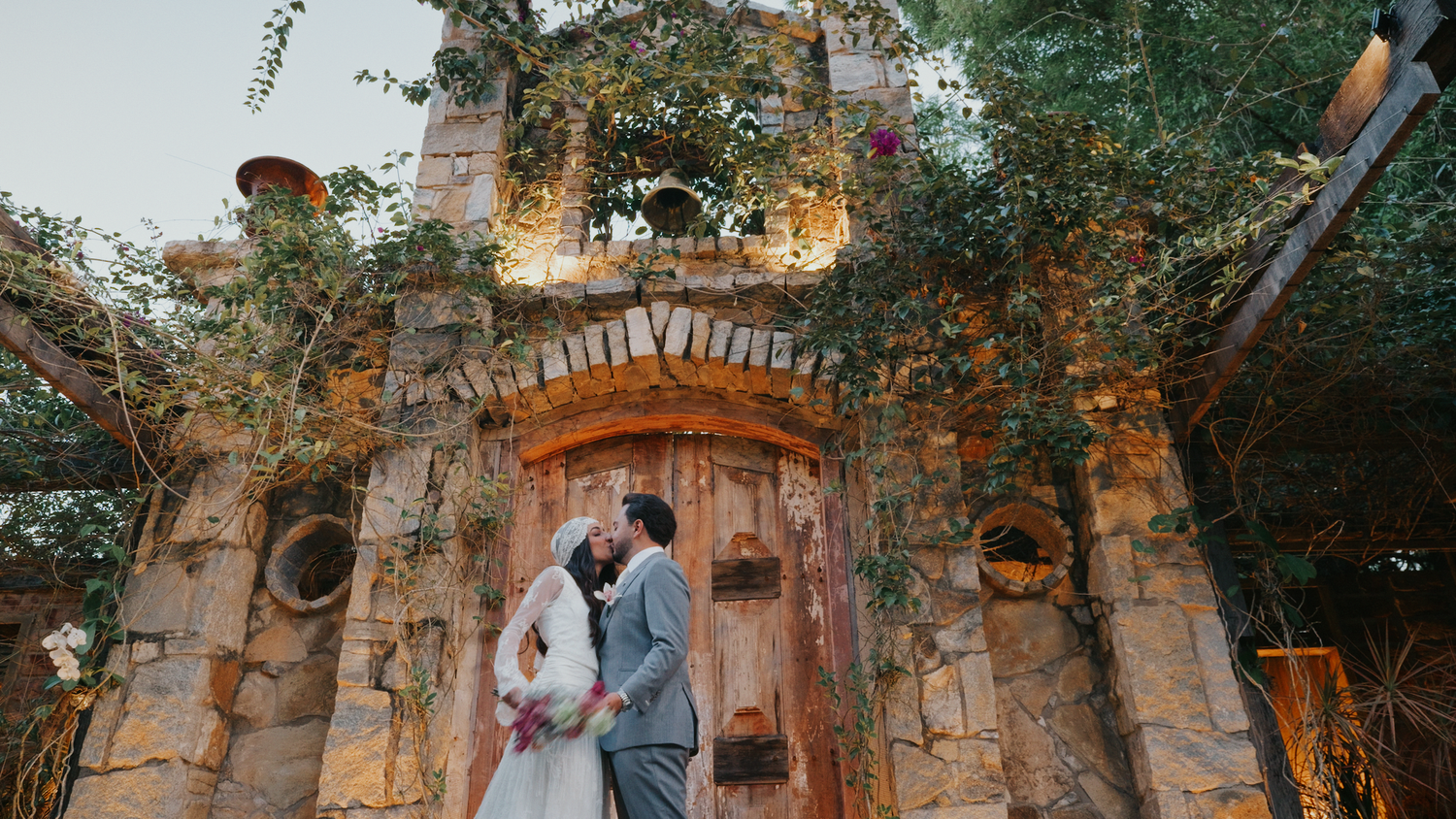 Bride and groom share a romantic kiss in front of a rustic stone archway at golden hour.